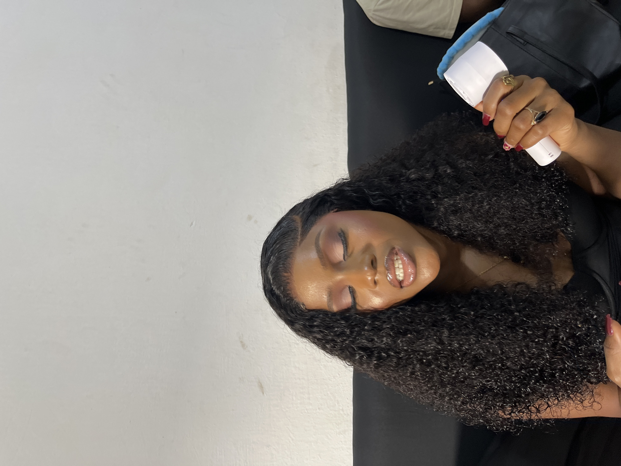 The scene is observed in an indoor setting, identified as Gura Top, Nigeria. A single adult Black female is centrally positioned, with her eyes closed and head slightly bowed. She presents with a full face of makeup, including pink/mauve eyeshadow and glossy lips with a slight gap revealing light-colored teeth. Her hair is dark, long, and curly, parted on the side, with a visible lace front. She wears a dark, possibly black, top. Her left hand is visible, featuring manicured nails with dark red polish, and she wears a gold-toned ring on her index finger and a gold-toned ring with a dark stone on her ring finger. She holds a white, cylindrical object, resembling a portable ring light, with a blue accent visible behind it. A dark bag or pouch is partially visible underneath the held object. She is seated on a black, upholstered surface, likely a sofa. The background consists of a plain, off-white wall exhibiting some smudges and discoloration. A partial view of another individual, clad in a light-colored (cream or beige) garment, is visible on the far right of the frame, adjacent to the primary subject.
