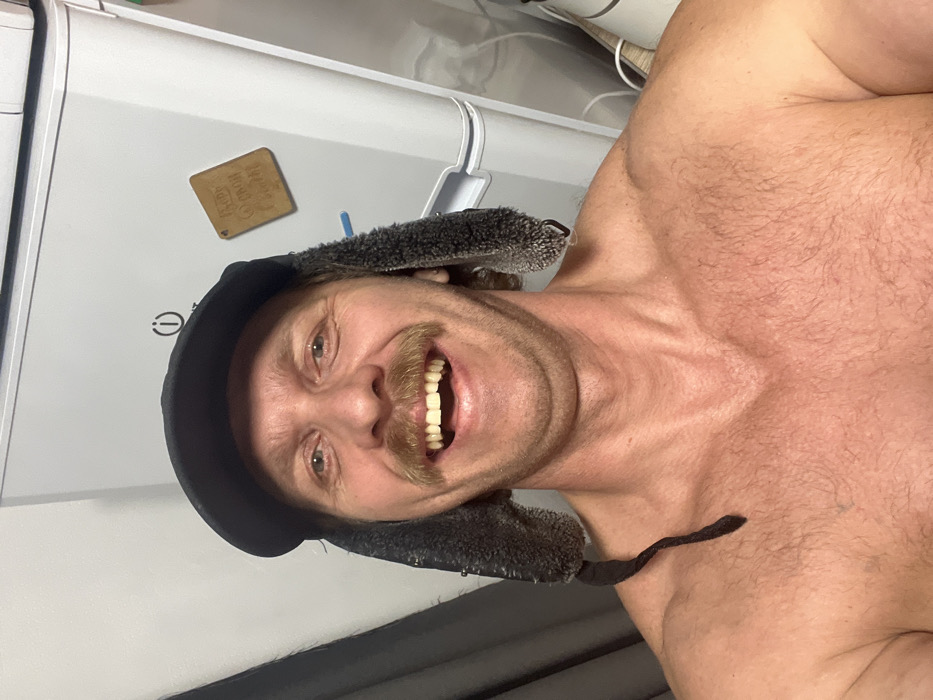 The image captures a close-up, chest-up selfie of a man, oriented vertically, showing him smiling broadly. He is shirtless, revealing a muscular physique with a covering of light body hair on his chest and shoulders. He has a prominent, well-groomed blonde or light brown mustache and light-colored eyes. His mouth is open in a wide smile, displaying his white teeth.
The man is wearing a dark-colored ushanka-style hat with fuzzy ear flaps that are pulled down, framing his face. The material of the ear flaps appears soft and textured.
The background consists of a white kitchen appliance, likely a refrigerator or freezer, against which the man is posing. On the surface of the appliance, a small, rectangular, light brown magnet or plaque is visible, featuring the word "HOME" in bold letters, and beneath it, the words "SWEET HOME" are partially visible. Below the magnet, a small electrical symbol (a circle with a vertical line through the top) is present on the appliance's surface. Above the appliance, some indistinct cables or wires are visible, suggesting an indoor setting.
The man's expression is one of cheerfulness and happiness, suggesting a lighthearted moment captured in a personal photo.