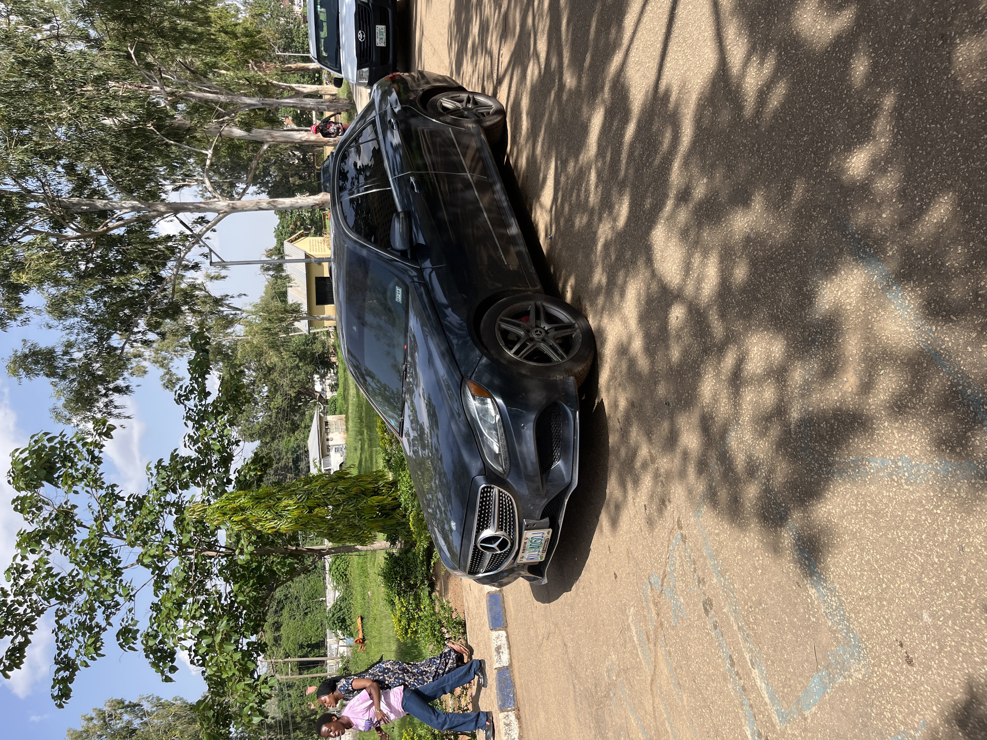 This media file shows a dark-colored Mercedes-Benz sedan parked on a paved surface, likely a road or parking lot. The car is angled towards the viewer, with its front end slightly closer. The scene is outdoors, with trees and greenery visible in the background. Sunlight casts dappled shadows from the trees onto the pavement and the car.
Two individuals are walking in front of the car, with their backs to the camera. One person is wearing a dark patterned dress and the other is in a pink shirt and blue jeans. They appear to be walking away from the car. In the background, a white pickup truck is parked behind the Mercedes-Benz. Buildings and more trees are visible further in the distance. The sky is blue with some scattered clouds.
The overall impression is a casual daytime scene. The presence of the parked cars and pedestrians suggests a public area, possibly a residential or commercial zone. There is no specific activity or event clearly depicted beyond people walking and cars being parked.
The license plate of the Mercedes-Benz is partially visible and appears to read "KUL406DU". The setting's context as Jos, Nigeria, is provided. The lighting suggests it is daytime, likely late morning or afternoon, given the angle and intensity of the shadows. The weather appears to be clear and sunny.