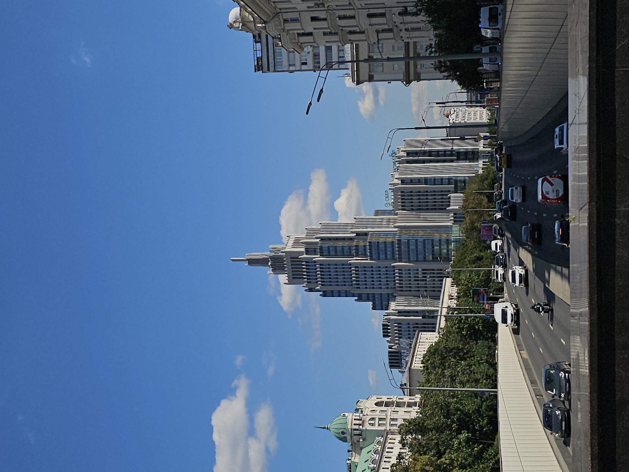This is a vertical, eye-level, medium shot of the Kotelnicheskaya Embankment Building, a Stalinist skyscraper located in Moscow, Russia. The building is centrally positioned and dominates the frame, its art deco architecture featuring a spire and setbacks. To the left of the skyscraper, a building with a green dome and a lighter facade is visible. The foreground is occupied by a multi-lane road with several cars and a motorcycle in motion, indicating traffic flow. The road is flanked by concrete walls, and trees line the embankment behind the road. The sky above is blue with scattered white clouds. The overall scene depicts a bustling urban environment on a clear day.