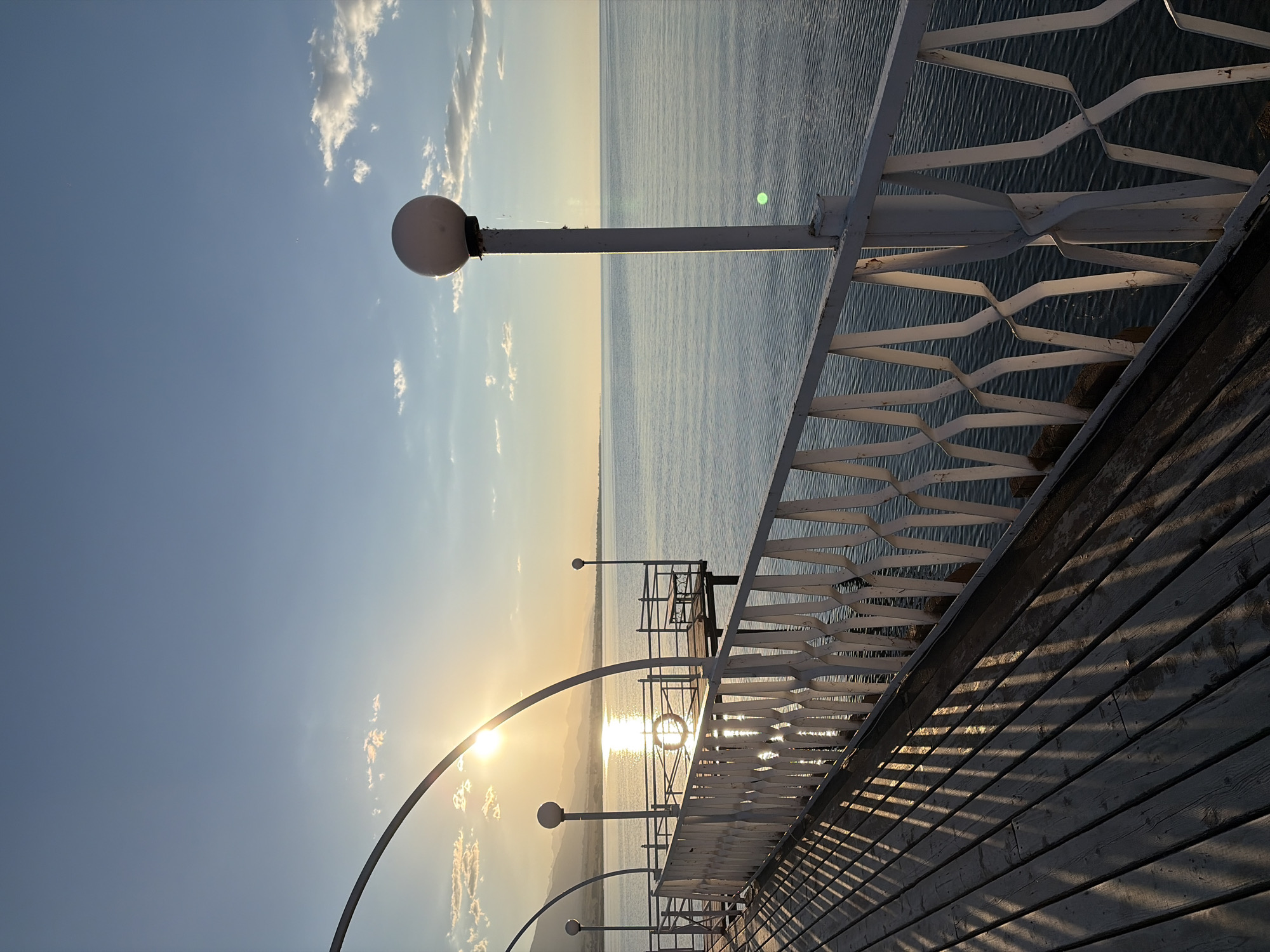 The image depicts a scenic view of a body of water, likely a lake or sea, under a clear sky with scattered clouds. The sun is setting or rising, casting a warm golden glow across the water and sky, creating a tranquil and serene atmosphere.
In the foreground, there is a wooden boardwalk with a white railing. The railing has an intricate, patterned design. A lamppost with a spherical light fixture stands on the boardwalk, near the railing. The sun's rays are reflecting off the water, creating a shimmering effect. Further along the boardwalk, there is a small pier with benches, suggesting a place for relaxation or observation. The shadows cast by the railing onto the wooden planks of the boardwalk are long and distinct, indicating the low angle of the sun.
The overall scene is peaceful and picturesque, likely a popular spot for enjoying the sunset or sunrise. The visual elements suggest a calm and beautiful natural setting, possibly a resort or park by the water. There is no visible text in the image.