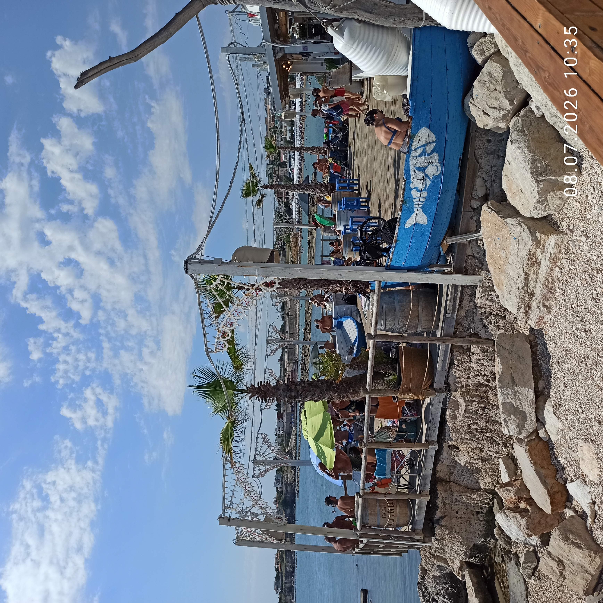In Trani, Italy, on July 8, 2026, at 10:35 AM, a waterfront establishment is depicted. The scene features a wooden deck elevated above rocky shoreline. The deck is populated with numerous individuals, many of whom appear to be patrons of the establishment. They are seated at tables, lounging, and standing. The establishment itself is decorated with white string lights and tropical plants, creating a relaxed, beachside atmosphere. A blue boat with a white fish design is prominently placed on the deck. In the background, the calm sea stretches towards a distant coastline dotted with buildings. The sky is a clear blue with scattered white clouds, indicating fair weather.