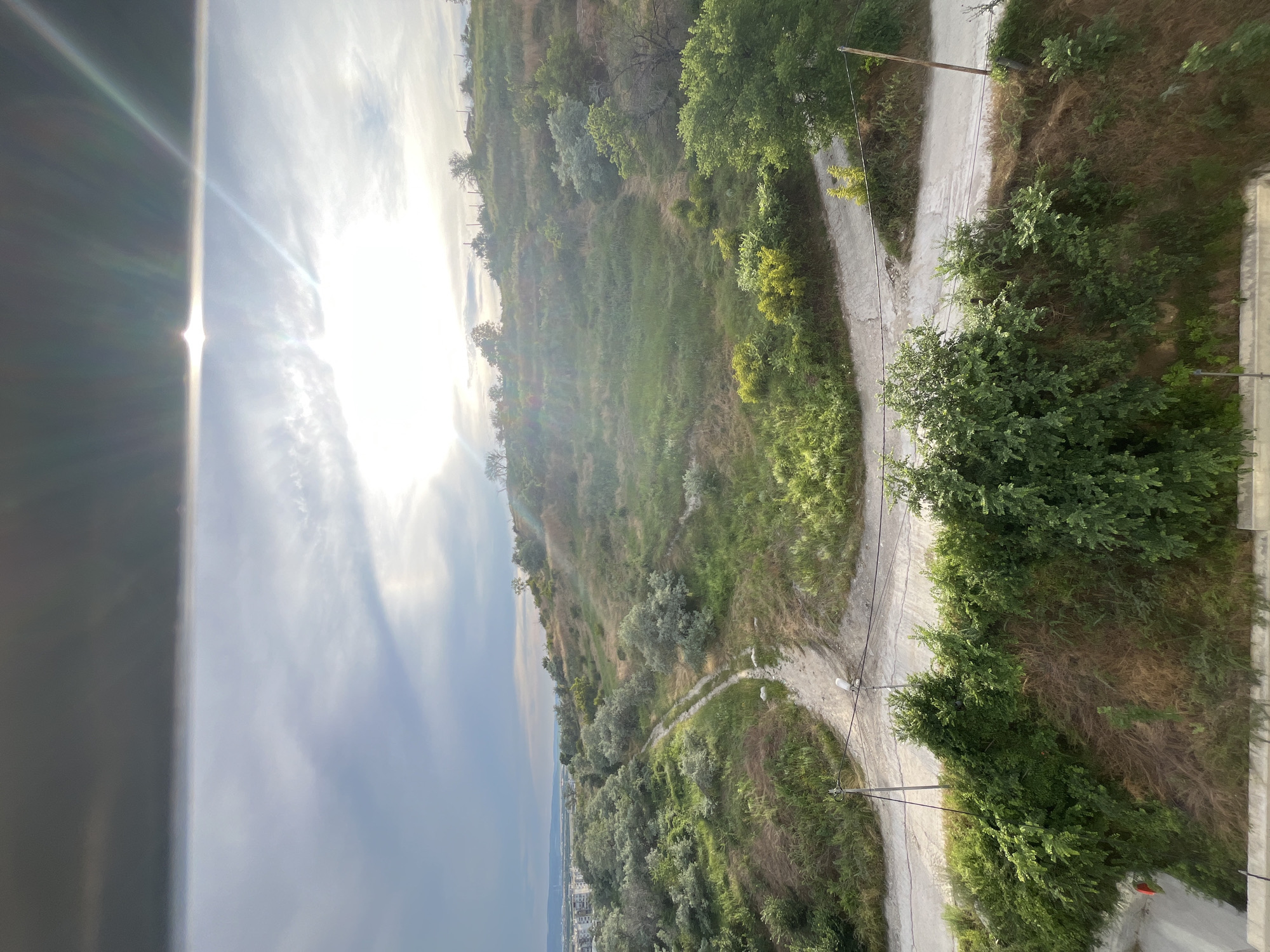 This is a photograph taken from an elevated position, looking out over a hilly, vegetated landscape with a road winding through it. The sun is visible in the upper right quadrant of the sky, casting bright light and lens flares across the scene. The sky itself is partly cloudy with a darker band of clouds across the top.
In the foreground, there is dense green foliage and a concrete structure, possibly a balcony or rooftop. A dirt or gravel road curves through the middle ground, splitting into two paths. The hills are covered in a mix of green grasses, bushes, and scattered trees, some of which appear to have silvery-grey foliage. In the distance, to the left, there are buildings that suggest an urban or suburban area, consistent with the outskirts of Varna, Bulgaria. The overall impression is a natural landscape juxtaposed with human development, captured during daylight with bright sunlight.
