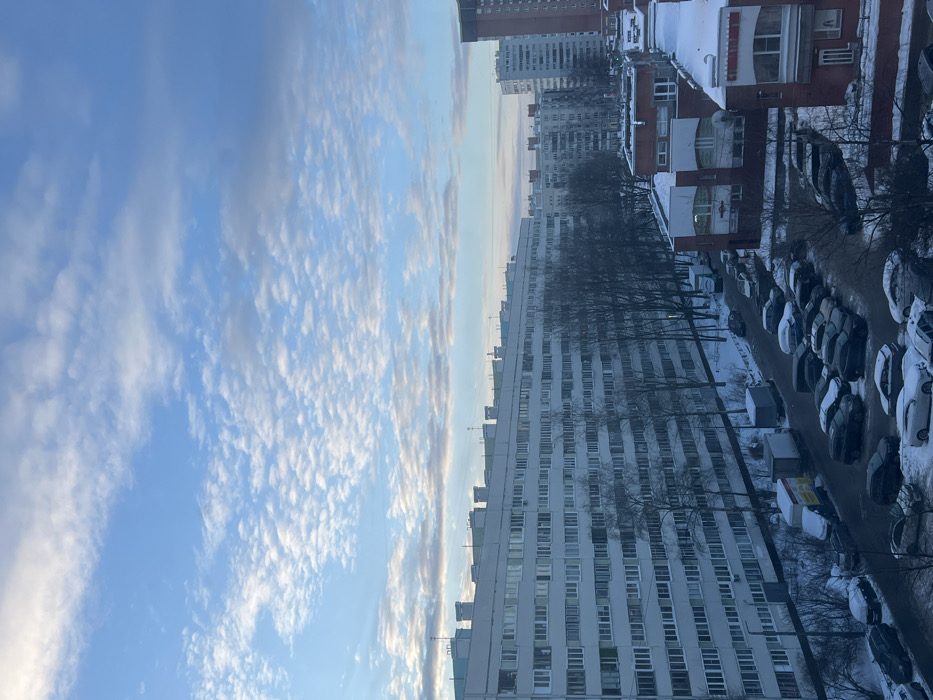 A vertical landscape shows a vibrant blue sky filled with patterned clouds. Below, a cityscape is visible, featuring densely packed apartment buildings. Snow lightly covers rooftops, parked cars, and ground surfaces near the buildings. Bare trees line the street, adding to the winter scene. The sky is bright, contrasting with the more muted tones of the urban setting.