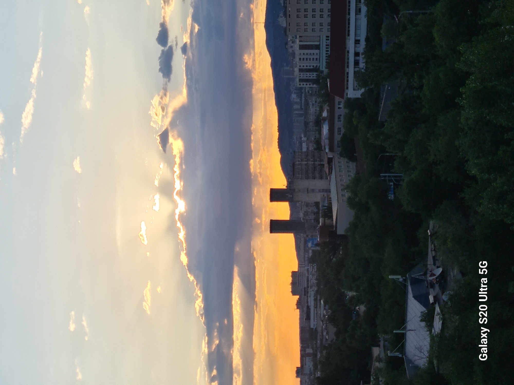 The image captures a cityscape at sunset in Almaty, Kazakhstan. The sun is partially obscured by dramatic, dark clouds that are illuminated by the setting sun, creating shafts of light. In the midground, a cluster of buildings forms the urban landscape, including two tall, dark cylindrical structures that stand out. Further in the background, a range of hills or mountains is visible against the vibrant orange and yellow hues of the sunset sky. The foreground is filled with dense green trees, suggesting a residential or park area within the city. A watermark at the bottom left corner reads "Galaxy S20 Ultra 5G".
