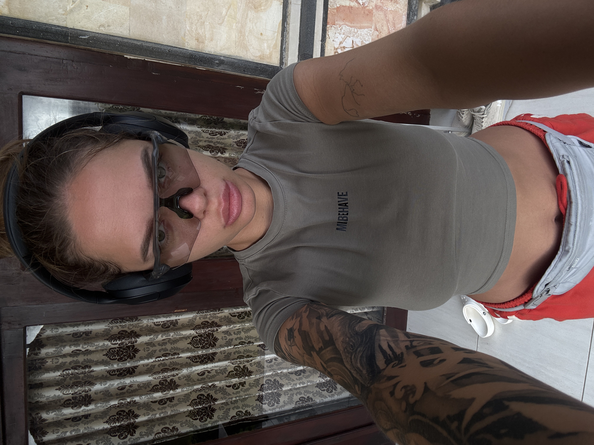 A selfie shot depicts a woman wearing headphones and sunglasses, positioned in front of a window in Unknown, Indonesia. The woman is wearing a short-sleeved gray t-shirt with "MIBBEHAVE" printed on it, along with red and gray shorts. The woman also has numerous tattoos on both arms. The window is covered by a patterned curtain. The background shows a tiled floor and a white shoe.