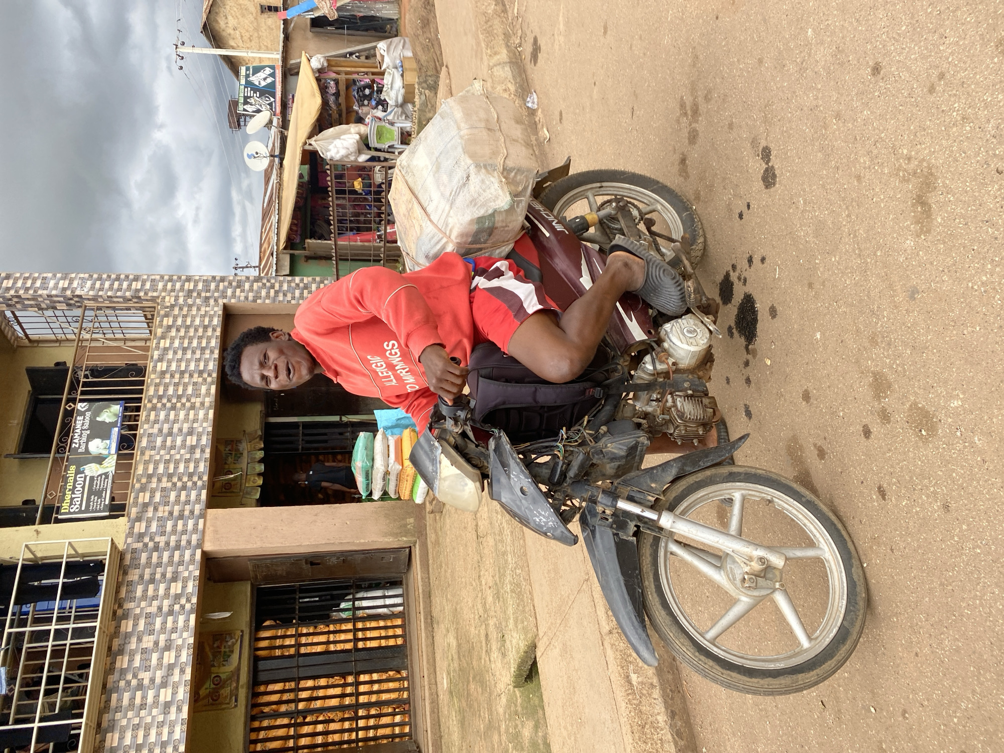 The image shows a person riding a motorcycle in the city of Bukuru, Nigeria. The person, wearing a red sweatshirt and red and white shorts, is looking towards the camera with an open mouth. They are seated on a dark-colored motorcycle, which is loaded with a large, bundled package strapped to the rear. A black backpack is visible on the motorcycle's seat behind the rider. The motorcycle appears to be somewhat worn, with exposed wiring on the front. The rider's feet are clad in dark grey slip-on shoes.
The background depicts a street scene in Bukuru. To the left, there is a building with a tiled facade and balconies. A sign for "Dhernails Saloon" and "ZAMANEES Barbering Saloon" is visible on the building's upper level. Large sacks of goods, possibly grain or other commodities, are stacked behind metal bars on the ground floor of this building. To the right of this building, there are market stalls with awnings and various goods displayed. Power lines and utility poles are visible in the distance against a cloudy sky. The ground is a dirt or asphalt surface with patches of oil stains.