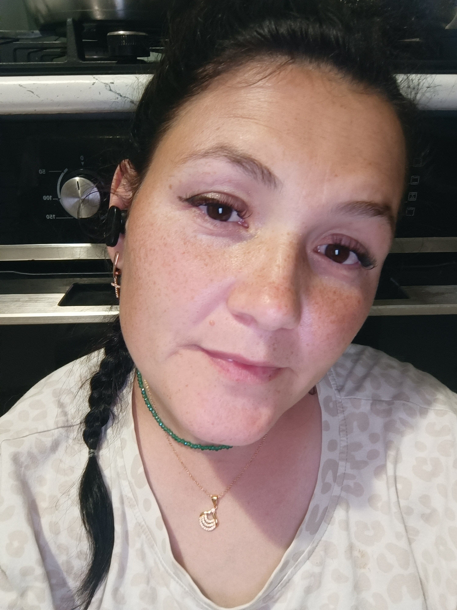 A close-up, selfie-style photograph shows a woman with dark hair pulled back and a braid in front of her left shoulder. She has fair skin with freckles, brown eyes, and is wearing makeup. She is wearing a light-colored t-shirt with a leopard print pattern. Around her neck, she has two necklaces, one with green beads and a gold pendant. She is also wearing small hoop earrings with a cross charm. In the background, a black stove top with silver knobs and a white speckled surface is visible. The image appears to have been taken indoors, likely in a kitchen, as suggested by the stove. The location in Chaikovsky, Russia, is not determinable from the visual cues present in the image.