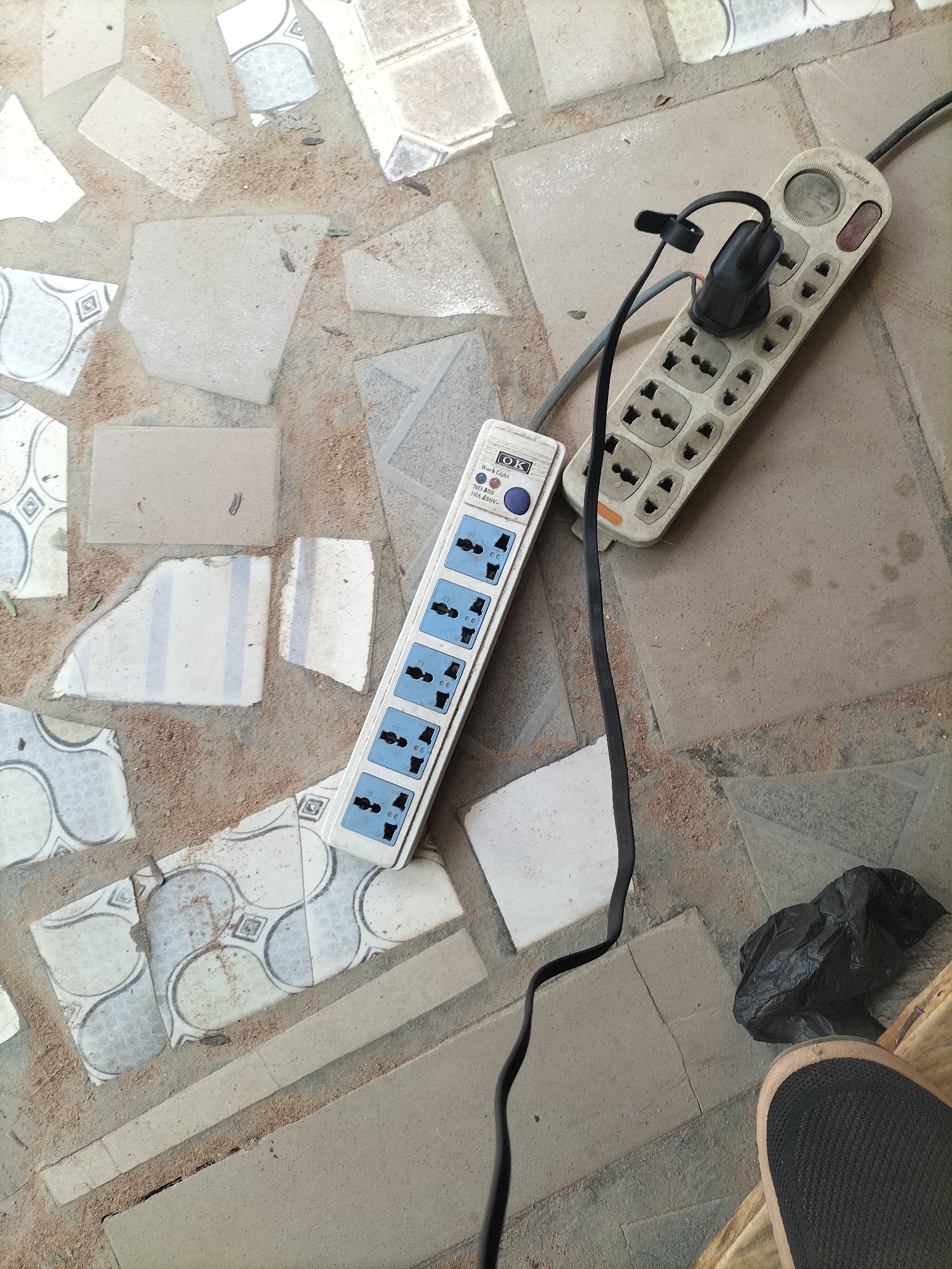 The image depicts an outdoor scene, likely a pathway or an unfinished floor, characterized by broken and uneven tiles interspersed with sandy soil. Two power strips are visible. One is a white, multi-outlet strip with blue receptacles, positioned diagonally across the frame. It has several outlets, each with a blue insert. The second power strip, also white, is placed to the upper right and features a different type of multi-socket design with a circular dial and a red indicator light at the top. A black electrical cord is connected to this second strip and extends downwards. A black plastic bag lies on the ground near the power strips. In the bottom right corner, a person's foot wearing a dark shoe with a tan accent is visible, suggesting a person is present at the scene. The overall environment appears somewhat neglected or under construction, consistent with a location in Jalingo, Nigeria.