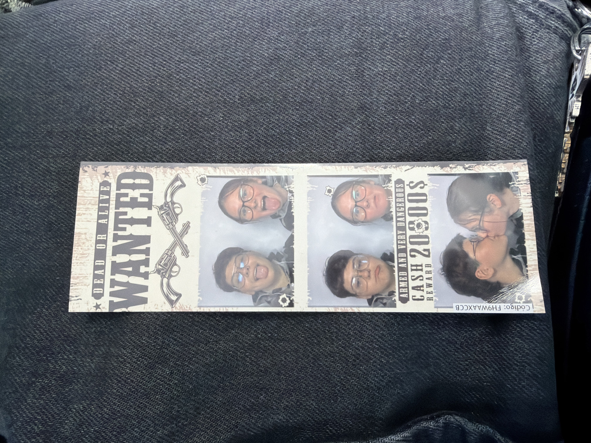 The image is a vertical photo booth strip. The strip has a "DEAD OR ALIVE WANTED" theme. The top panel shows two people sticking their tongues out. The second panel also shows two people, one with a serious expression and the other with their tongue out. The third panel shows two people with surprised expressions. The bottom panel shows two people kissing. The photo strip has a code on the left side: "Codigo: FH9WAAXCCB". The background of the photo strip is a denim texture. The photo strip is placed on a dark denim fabric. There are no visible landmarks or indicators that place this image in the city of Sorø, Denmark.