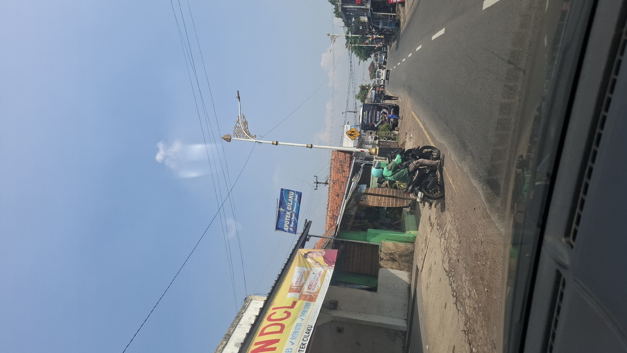 The image depicts a street scene in Cilaku, Indonesia, likely on a clear, sunny day. On the left side of the road, a building with a white facade features a prominent yellow and red banner that reads "NDCL." Below this banner, another banner displays "APOTEK CILAKU." A green awning extends from the building, and a small, open-fronted shop with a brick facade is situated next to it. A person wearing a green jacket and helmet is seated on a black motorcycle parked on the side of the road, facing away from the camera. The road itself is paved and marked with white lines. Further down the road, several vehicles, including a bus and other cars, are visible, traveling in the same direction as the camera's perspective. A pedestrian crossing sign with a yellow background and black silhouette of children is posted on the right side of the road. Decorative streetlights with ornate gold and white designs line the street. The environment appears to be a mix of commercial establishments and residential or semi-rural structures.