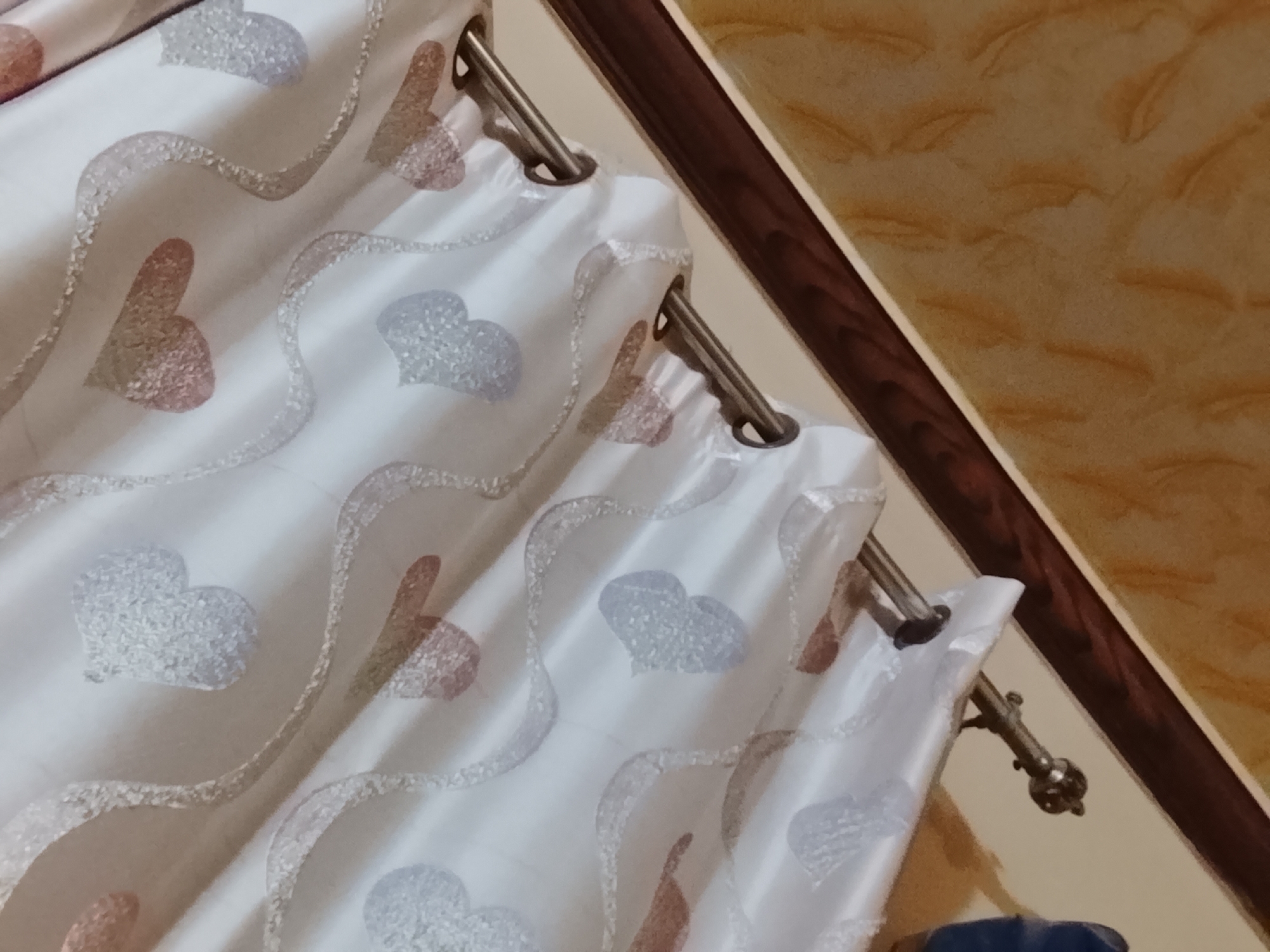 A close-up, slightly angled shot focuses on a section of white curtains with a heart pattern hanging from a curtain rod. The hearts alternate in color, appearing as a light pink or a light blueish-gray. The curtain rod is metallic and mounted against a wall painted in a marbled yellow tone, framed by a dark brown wooden trim. The bottom right corner shows a hint of what appears to be a dark blue object. Given the request for location information, this scene is likely located in a private residence in Multan, Pakistan, based on the cultural context suggested by the image of household items.