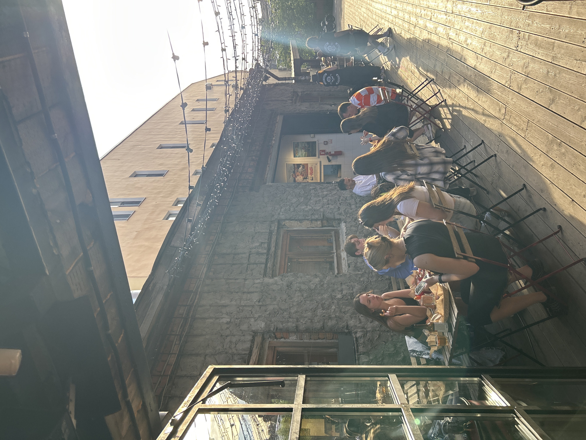 This image captures an outdoor dining scene at a cafe or bar in Saint Petersburg, Russia, during the late afternoon or early evening with strong, directional sunlight. The foreground features a large, dark-framed open window reflecting the outdoor setting. The ground is a light brown wooden deck, with visible planks and gaps, extending across the entire lower half of the frame.
Approximately 10-12 individuals, mostly young to middle-aged adults, are seated at several foldable tables with metal frames and wooden tops, accompanied by matching foldable chairs with wooden slats. On the left, two women face each other; one, with blonde hair in a bun and a black sleeveless top, is intently looking at a smartphone. The other, with long dark hair and a black tank top, holds a snack to her mouth. Other individuals are seated around them and at additional tables further back, eating, drinking from bottles and glasses, and engaging in conversation. Two men are visible at a table in the mid-ground, one in a white t-shirt and another in an orange and white striped t-shirt.
In the background, two individuals wearing dark shirts with white circular logos on their backs are standing or walking towards the right. A motorcycle is partially visible behind them. The main structure on the left is a single-story building with a rough, textured grey stone or concrete exterior. It features two wooden-framed windows with dark panes and a larger open bay housing two posters. Decorative string lights with numerous small bulbs are strung overhead, extending from this building across the outdoor area. Behind the cafe, a multi-story building with a light brown facade and multiple rectangular windows is brightly illuminated by the sun, which causes a bright flare on the right side of the image and overexposure in the sky. Long shadows of people and furniture are cast across the wooden deck due to the low sun angle.