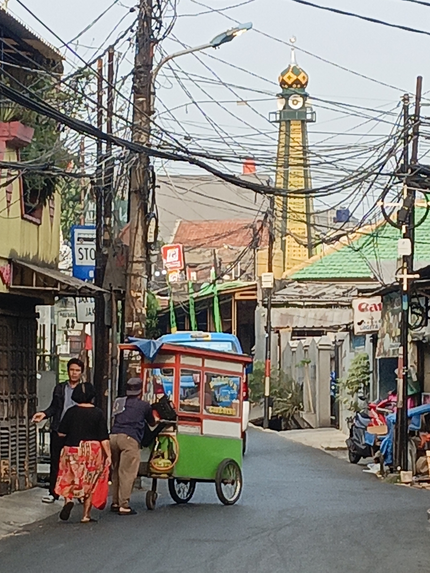 In a narrow street in the Duren Sawit subdistrict of East Jakarta, Indonesia, a street scene unfolds beneath a chaotic tangle of overhead wires. A colorful food cart, green and red with a blue awning, sits on the asphalt road. Two men and a woman are near the cart, possibly interacting with the vendor or the cart itself. One man, wearing a dark jacket, stands facing the cart, while another man, in a blue shirt and grey pants, is behind the cart, appearing to be working or tending to it. A woman in a brightly patterned red skirt and black top walks away from the cart. In the background, a minaret of a mosque with a distinctive patterned dome rises above the rooftops, its architectural details visible despite the network of wires. Buildings lining the street are a mix of yellow, green, and reddish-brown, with some featuring awnings and visible windows. A street sign with "STO" in blue and white is mounted on a pole, and other signs are present on buildings. Motorcycles are parked on the right side of the street. The scene suggests a bustling urban environment, common in Jakarta's residential areas.