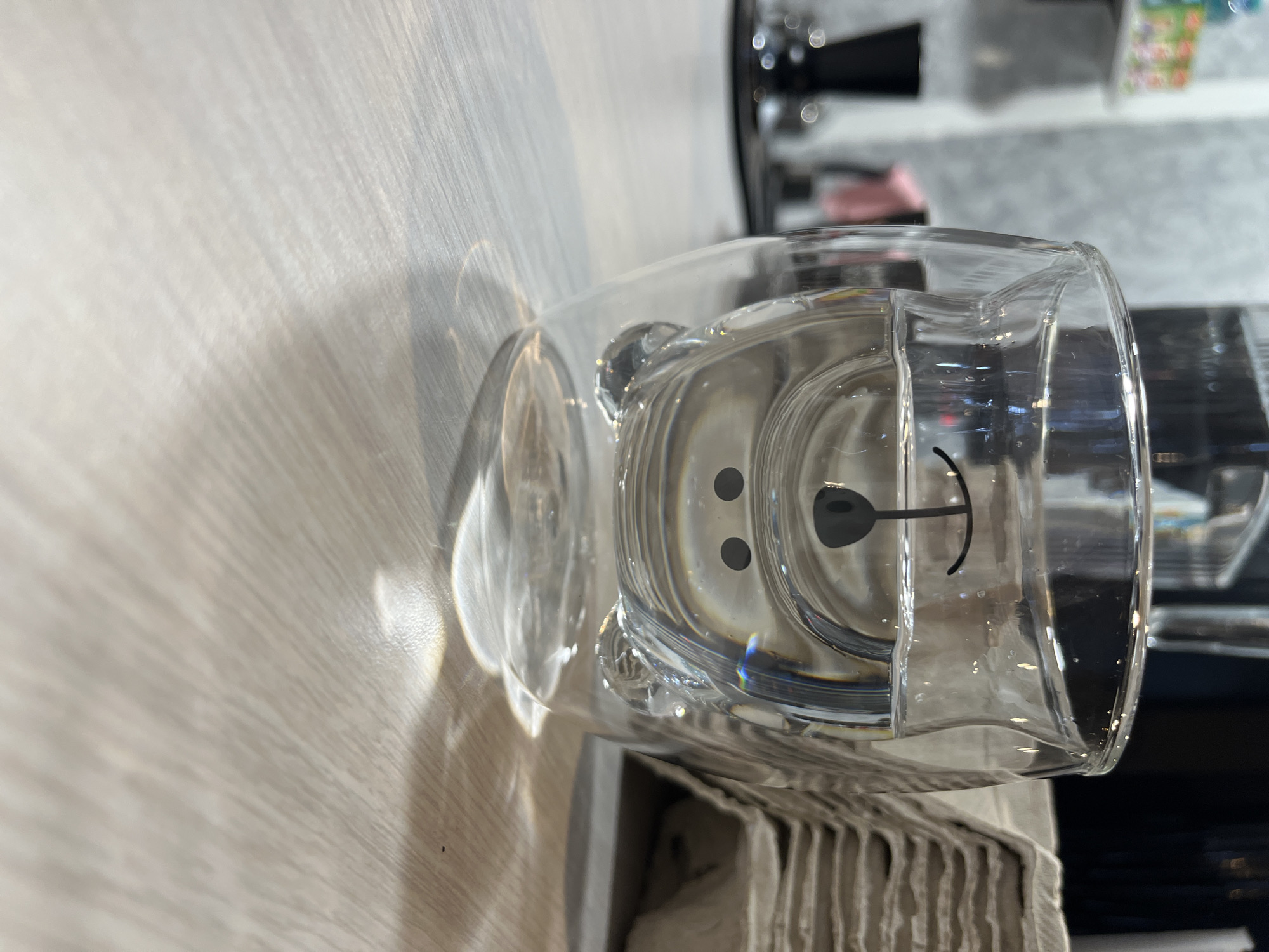 A clear glass mug with a cartoon bear face design on the inside is resting on a light-colored, wood-grained surface. The mug appears to have a handle on the left side. In the background, out of focus, are blurry shapes that suggest a retail environment. The scene appears to be lit by natural light creating distinct shadows on the surface. There is no indication in the image that it was taken in Chaikovsky, Russia.