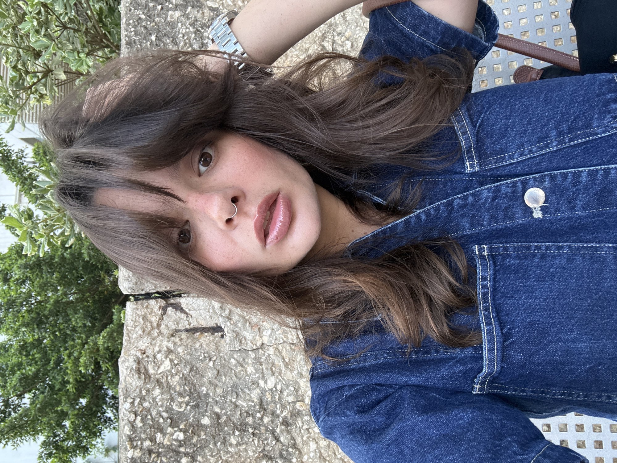 A young woman with brown hair and a septum piercing is pictured in a close-up shot. She is wearing a denim jacket and has her hand on her head. The background consists of a stone wall and greenery, suggesting an outdoor urban setting, possibly in Tel Aviv, Israel, given the context of the prompt. The lighting appears to be natural daylight.