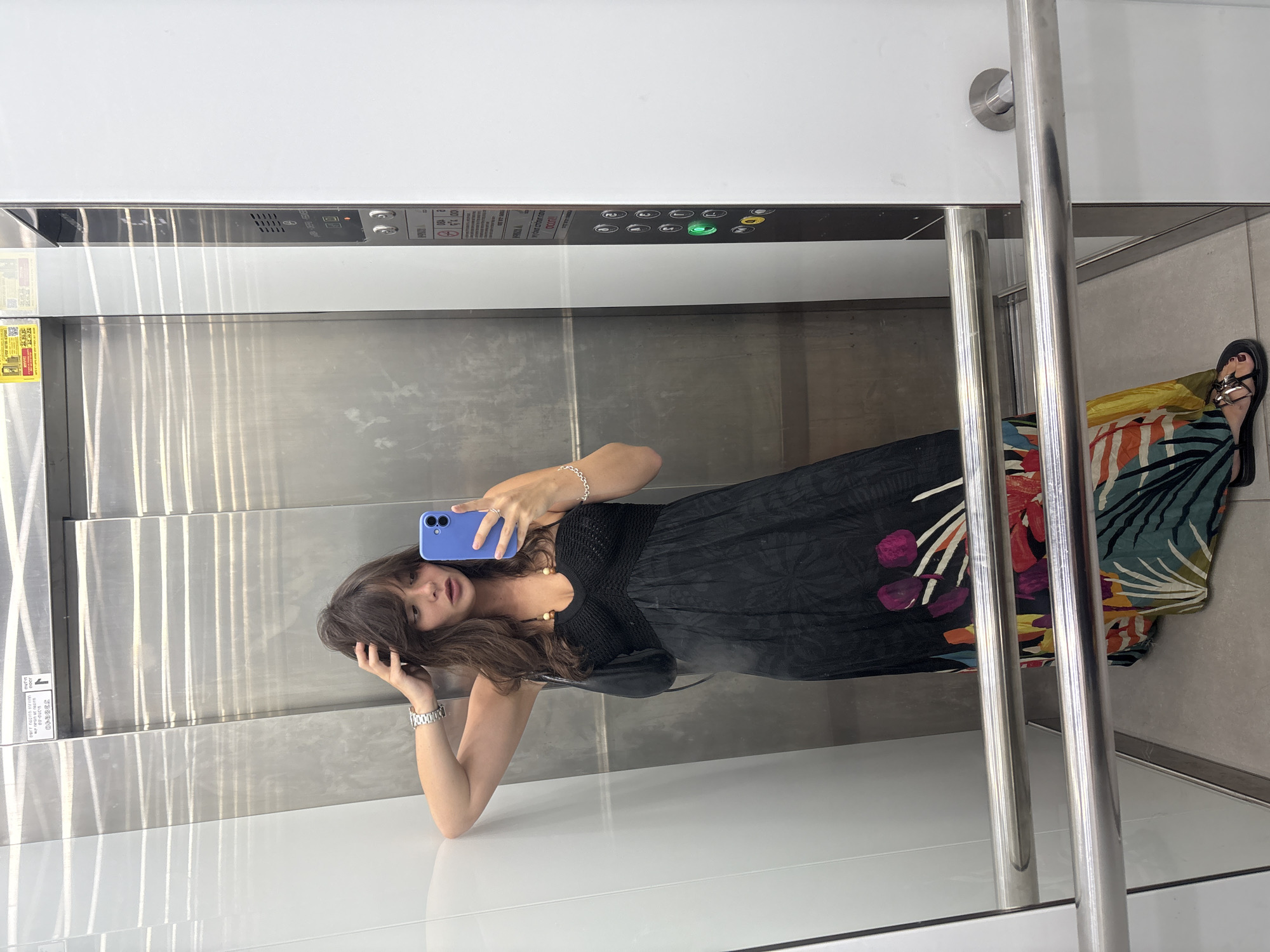 A young woman with brown hair is taking a mirror selfie in an elevator. She is wearing a black sleeveless maxi dress with a colorful floral and tropical print at the hem, a silver wristwatch, and a silver bracelet. Her right hand is in her hair, and she is holding a blue smartphone in her left hand. She is also wearing black sandals. The elevator has brushed metal walls and a white panel with buttons on the right side. A metal railing is visible in the foreground. The setting appears to be an elevator in Tel-Aviv, Israel, indicated by the elevator button panel which includes Hebrew text and symbols common in Israeli public spaces.