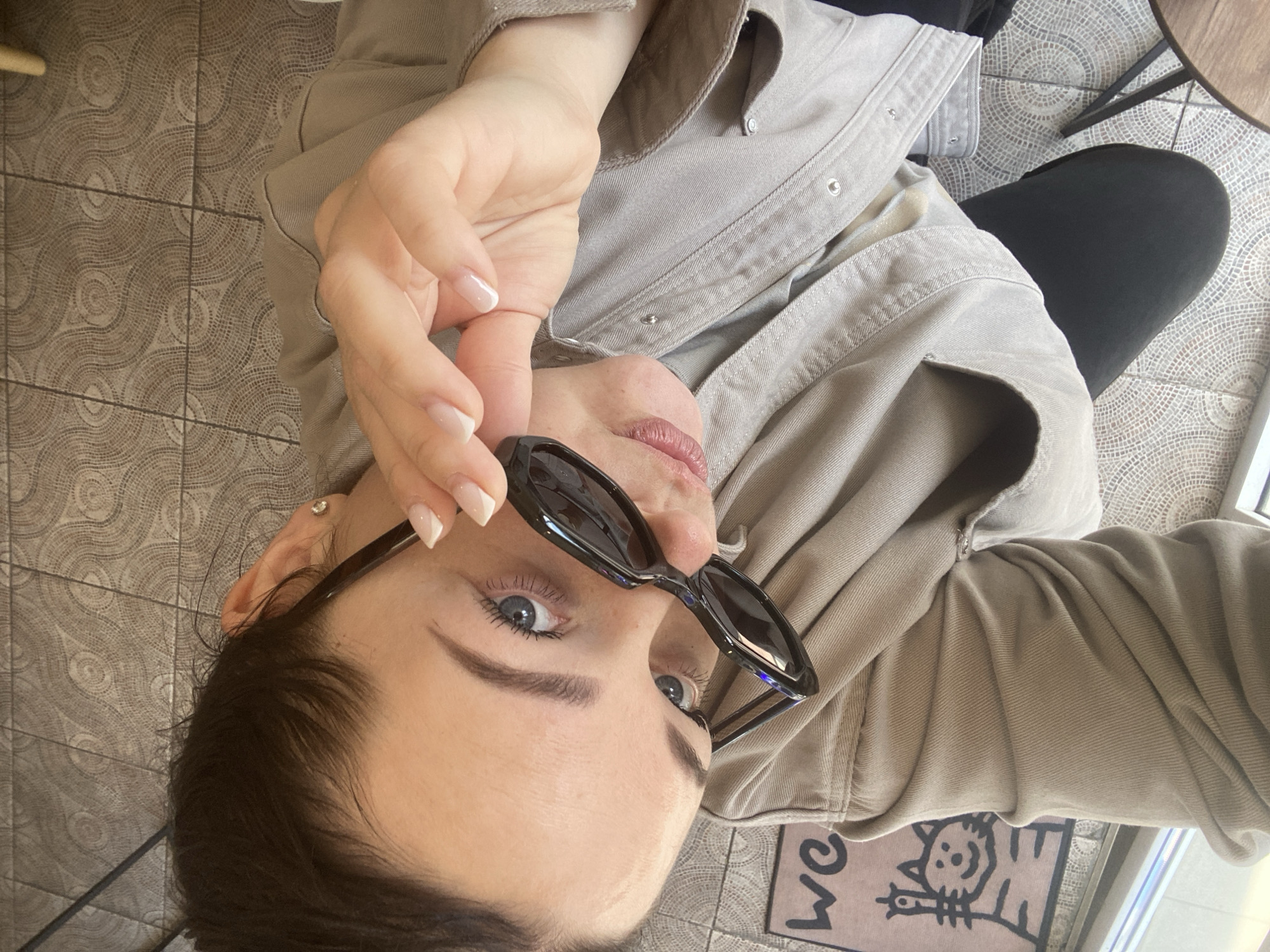 A low-angle, rotated close-up captures a young adult female looking directly at the camera over her sunglasses. She has light blue/grey eyes, dark eyebrows, and dark hair, with a small diamond stud earring visible in her left ear. Her hand, adorned with nails manicured with white french tips, holds down a pair of black, angular sunglasses.
She is wearing a light beige or khaki-colored jacket or shirt, possibly denim or canvas, with silver snap buttons. A dark top is visible underneath, and she wears dark pants or leggings. The setting is indoors, likely a cafe or home entrance in Sochi, Russia. The floor is tiled with square tiles, each featuring a repetitive, intricate mosaic-like circular pattern in shades of brown and beige. A small section of a light-colored doormat is visible in the bottom right, displaying the text "we" and a black outline illustration of a smiling cat. The image has a casual and direct feel, with the subject engaging the viewer.