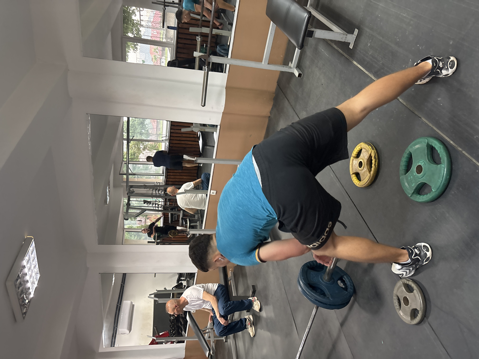 The image shows the interior of a gym in Zlatograd, Bulgaria. A young man is performing a barbell row with a blue weight plate on the barbell. To his right and in the background, there are several other weight plates of varying colors (green, yellow, gray). The man is wearing a blue t-shirt, black shorts, and black and white sneakers. In the background, another man in a white shirt and blue pants is seated on a bench. Mirrored walls on the left side of the frame reflect the gym interior, including other people and equipment. A weight bench is visible at the top of the frame. The floor is black and made of rubber.