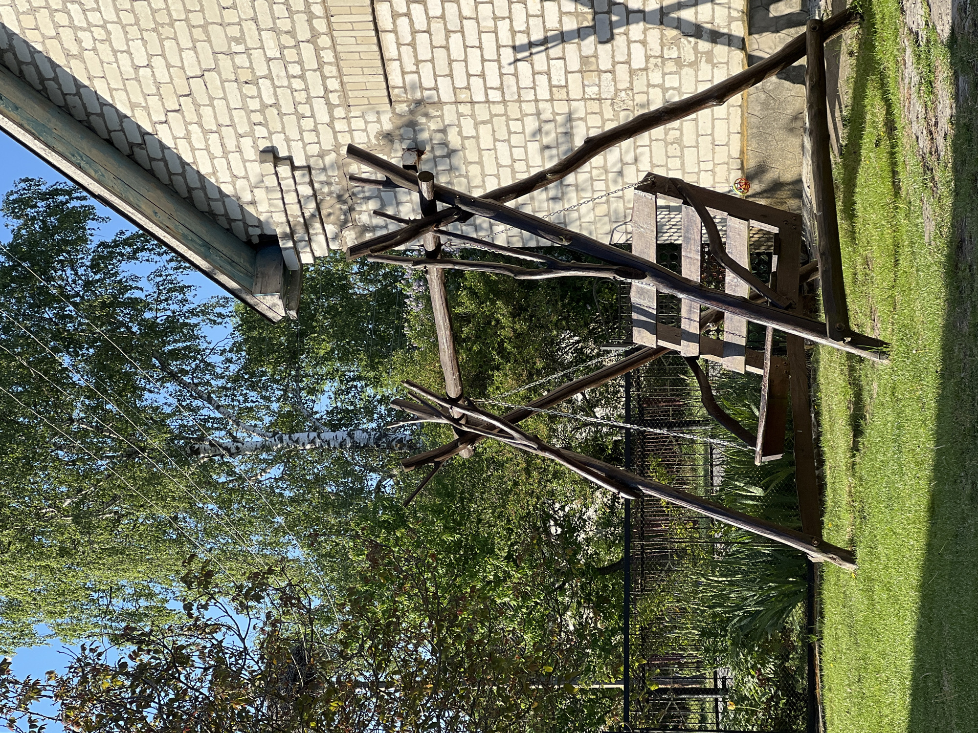 The image presents a scene in what appears to be a backyard or garden setting. A wooden swing set dominates the foreground, constructed from rough-hewn timber and chains. It is located on a patch of green lawn. A black, metal fence marks a boundary behind the swing. Beyond the fence, lush greenery, including trees and shrubs, fills the background. Part of a light-colored brick building is visible in the upper portion of the image. Overhead electrical wires are noticeable cutting across a portion of the sky. Although the exact location is specified as Vertelka, Ukraine, this can not be verified within the visual scene. No people are visible in the scene.