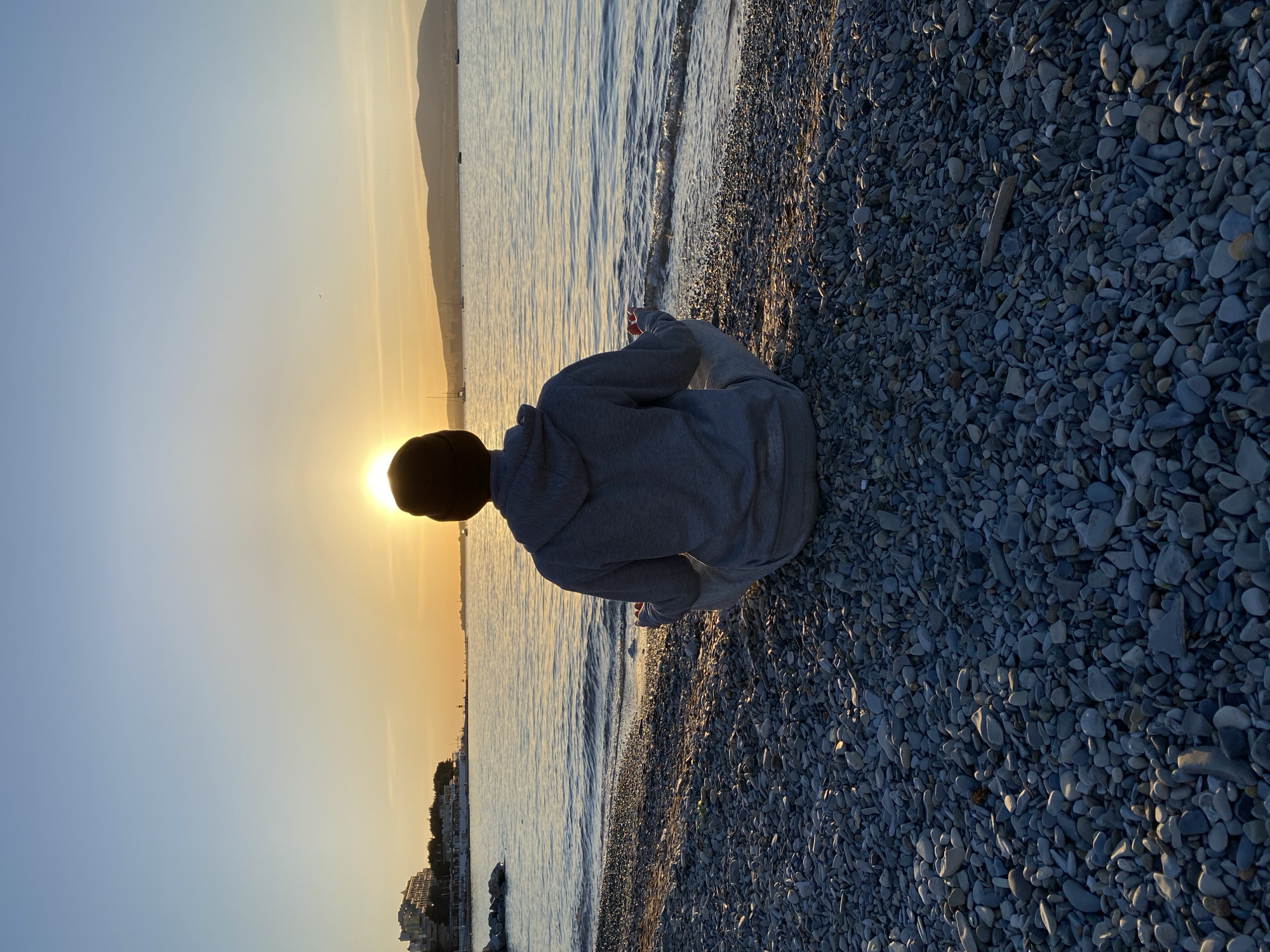 This image captures a serene moment on a pebble beach in Gelendzhik, Russia, during either sunrise or sunset. A person, viewed from behind, sits peacefully on the shore, facing a calm body of water. They are wearing a grey hooded sweatshirt and a dark cap, their silhouette partially obscuring the bright, low-lying sun on the horizon.
The sun is a powerful, radiant orb, casting a warm golden glow across the water's surface and reflecting a soft light onto parts of the pebble beach. The beach comprises countless smooth, grey and dark-toned pebbles, some highlighted by the sun's gleam. Gentle ripples mark the edge where the water meets the shore.
In the distance, across the expansive water, a hazy line of mountains or hills stretches along the horizon. Along the coastline, faint outlines of buildings and structures are visible, characteristic of a coastal town. Several small boats or yachts can be discerned on the water in the middle distance. The sky above transitions from a clear light blue to a warm, soft orange-yellow near the sun, indicating the early morning or late evening. The scene evokes a sense of calm and contemplation. There is no visible text in the image.