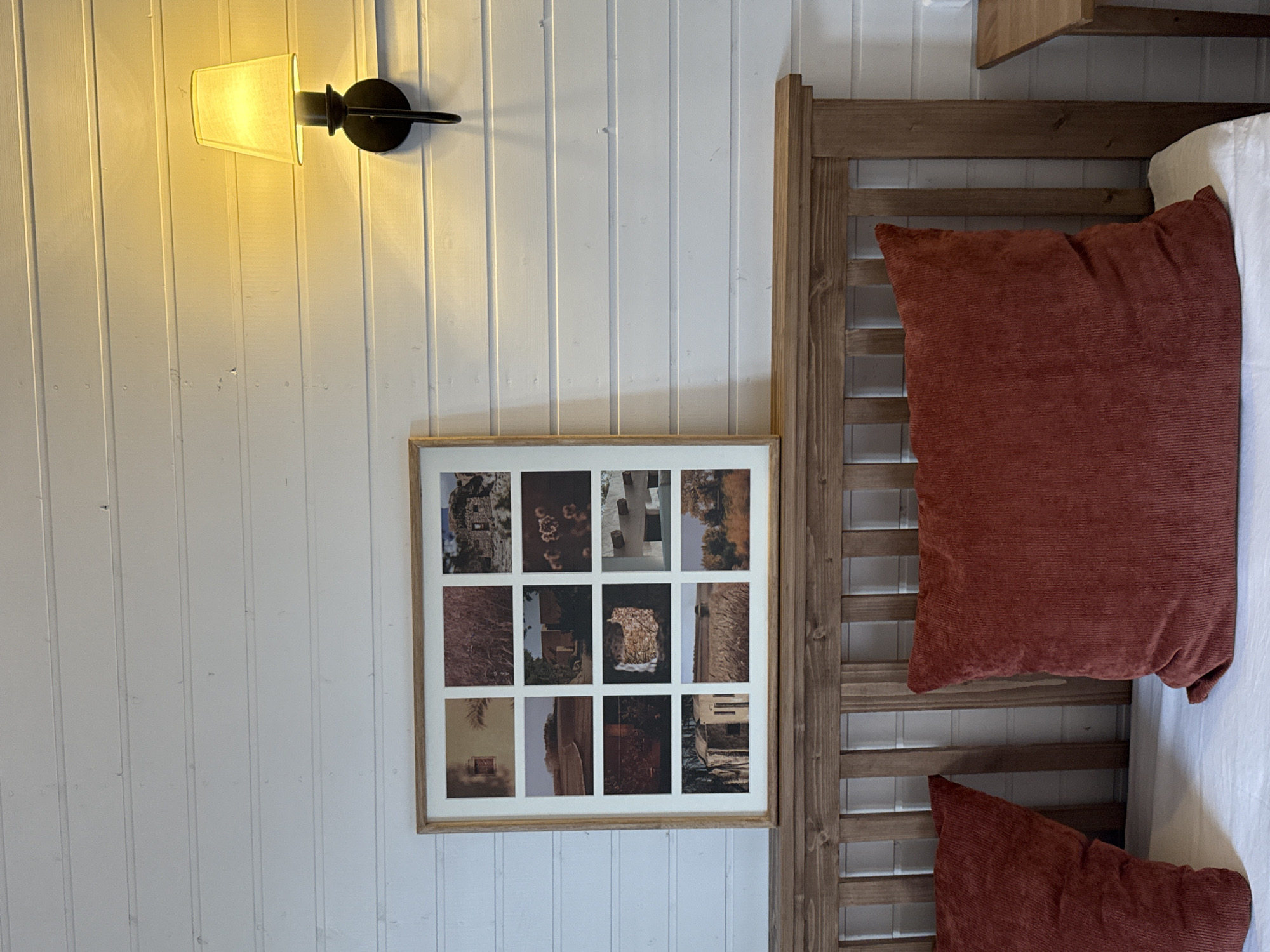 This is an indoor photograph capturing a cozy bedroom interior in Starokorsunskaya, Russia. The dominant feature is a white wooden paneled wall, which runs vertically through the left and central parts of the frame.
Mounted on the wall, in the upper left, is a black metal wall sconce with a soft yellow fabric lampshade, emitting a warm, artificial light. Below the lamp and slightly to the right, a rectangular wooden-framed picture is displayed. This frame contains a grid of twelve smaller photographs, arranged in three rows and four columns, depicting various architectural details and elements, possibly from a historical or rustic location, in muted, earthy tones.
To the right of the image, a wooden bed frame or headboard is visible, featuring horizontal wooden slats. Against this headboard are two large, rectangular pillows covered in a rust-red or burnt orange corduroy-like fabric. A white sheet or duvet covers the bed mattress, partially visible. Above the headboard, a small wooden shelf or beam is attached to the wall.
No specific activity is taking place in the static scene. The lighting suggests it is either evening or that the room relies solely on artificial light. There is no visible text in the image.