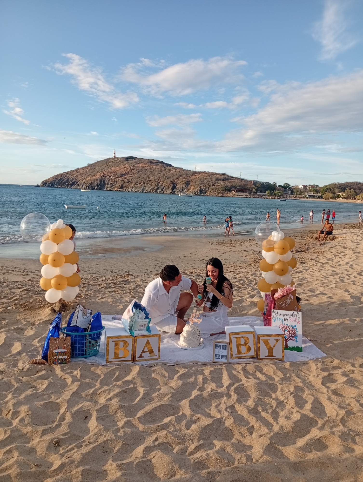 A medium shot captures a gender reveal event on a sandy beach against a backdrop of the sea and a land formation in the distance, potentially in Pedro González, Venezuela. In the foreground, two individuals are seated on a white cloth laid out on the sand, surrounded by decorations.
To the left, a light-skinned man in a white button-down shirt and white shorts is holding a microphone or similar device. A dark-haired woman in a white dress is to his right, also holding a similar device. Between them is a two-tiered cake on a white cake stand.
Behind the couple, gold and white balloons arranged in columns are on either side of the white cloth, with gifts, cards, and a blue basket scattered around. In front of them are large wooden blocks spelling out "BABY" in gold lettering. The sign "Bienvenidos Boy or God?" is also visible.
In the background, people are enjoying the beach. The sea is visible between the beachgoers and a sizable land formation with a structure at its peak. The sky is a clear blue with scattered white clouds. The overall scene suggests a celebratory and public event in a beach setting.
