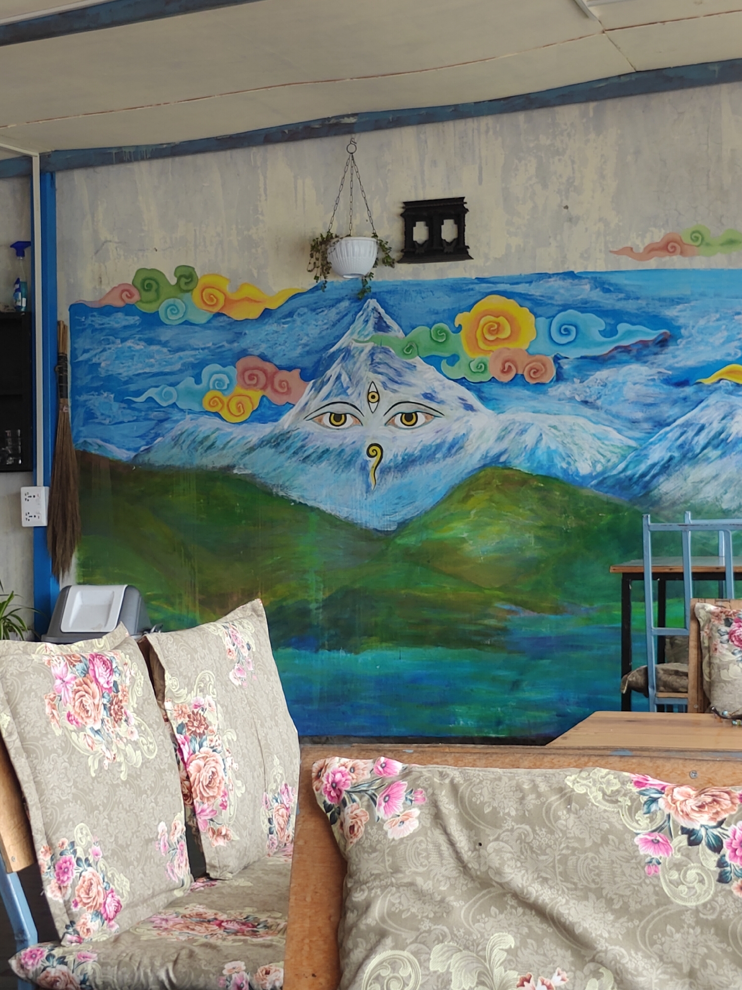 The image depicts a mural on a wall in Pokhara, Nepal, featuring a majestic mountain with a third eye prominently placed in its center, surrounded by colorful clouds and blue skies. Below the mountain, rolling green hills transition into a serene blue body of water. In the foreground, a couch or bench is visible, adorned with floral-patterned cushions. A wooden table with a blue chair is positioned to the right of the couch. The setting appears to be an outdoor or semi-outdoor area, possibly a cafe or guesthouse, with a broom leaning against the wall to the left of the mural. The overall impression is one of artistic expression combined with a relaxed seating area.