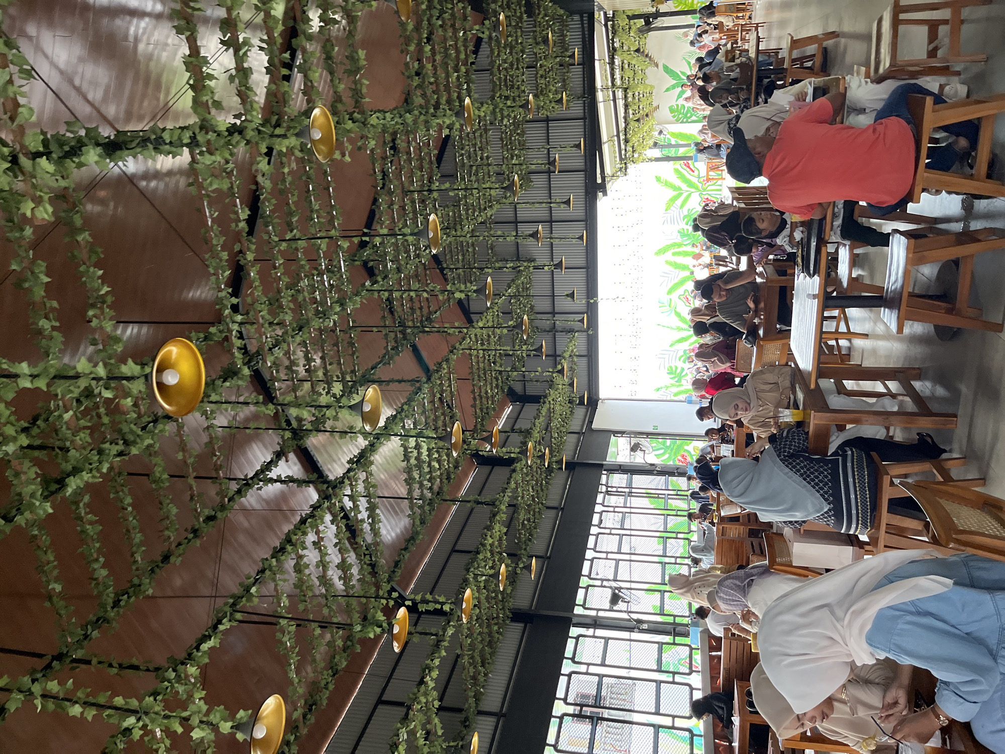 The image depicts an indoor dining area, likely a cafe or restaurant, located in Lhokseumawe, Indonesia. The overhead ceiling features a network of black structural elements from which numerous artificial green leafy vines are draped in a grid pattern. Interspersed among the vines are numerous inverted yellow bowl-shaped pendant lights, providing illumination. The ceiling itself is a mix of brown paneled sections and corrugated metal towards the right side.
The establishment is populated by approximately 30-40 identifiable individuals, comprising men, women, and children. Many of the women are wearing hijabs. Patrons are seated at wooden tables with matching wooden chairs and stools. Actions include eating, drinking from glasses (some containing orange beverages), engaging in conversation, and using mobile phones. For example, in the immediate foreground on the left, a woman in a light blue long-sleeved top and a white hijab looks down, while another woman in a beige top and light hijab is seated beside her. In the mid-ground right, a man in a red t-shirt and a dark baseball cap is seated with his back to the viewer, next to a child and a woman in a white top and hijab.
The left wall of the space appears to be composed of large, rectangular window panes or open grilles, allowing natural light into the area. The far wall features a large mural depicting stylized green and white tropical leaves. The floor is dark and non-reflective. The overall environment is one of a busy, casual dining setting.