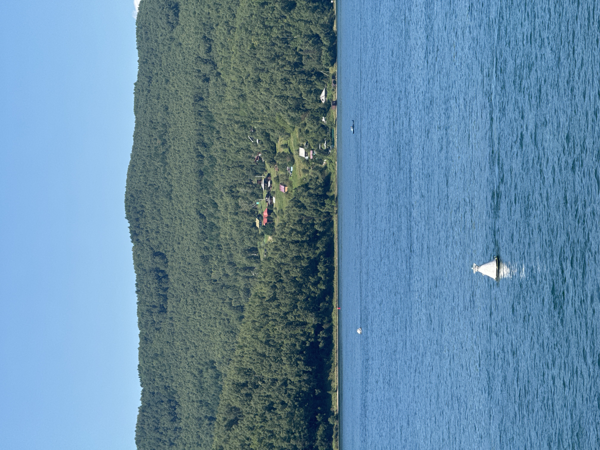 A wide shot captures a tranquil, deep blue body of water, likely Lake Baikal, under a clear, bright blue sky. A single, white, conical buoy floats prominently in the foreground, its reflection shimmering on the water's surface. In the mid-ground, a small, dark-hulled boat is visible on the water. The background is dominated by a densely forested, undulating green hillside. Scattered across the hillside are several small, red-roofed houses, indicative of a settlement. The scene conveys a sense of peaceful natural beauty and remote habitation. This location is the village of Listvyanka, Russia.