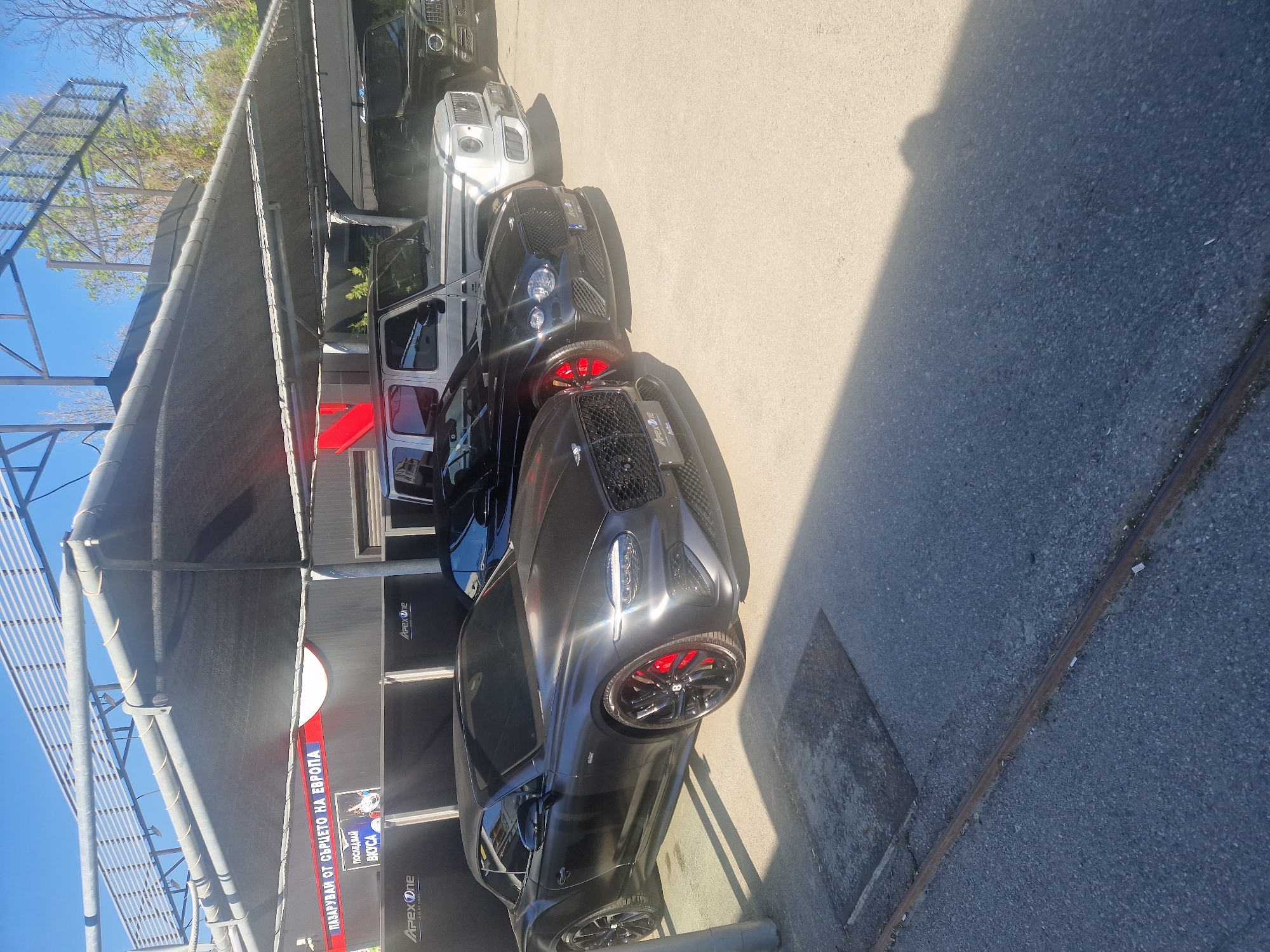 The image, oriented vertically, captures an outdoor scene featuring several luxury SUVs parked under a shaded structure. In the foreground, a dark grey/black Bentley Bentayga, featuring a wide-body kit, black multi-spoke wheels, and prominent red brake calipers, is parked. Behind it, a black Mercedes-Benz G-Wagen is partially visible, followed by a white G-Wagen, and another dark G-Wagen further down the line.
The vehicles are situated on a concrete and asphalt surface. Overhead, a metallic framework supports a grey tarp-like covering, providing shade from the bright daytime sun, which casts distinct shadows. The sky in the background is clear and blue, with some trees visible.
The setting appears to be a car service facility, possibly a high-end car wash or detailing shop. This is suggested by the presence of multiple luxury vehicles and visible signage. "ApexOne" branding is visible in several places on the structure and potentially on a car's license plate frame. A large red sign with Bulgarian text reads "ПАЗАРУВАЙ ОТ СЪРЦЕТО НА ЕВРОПА" (SHOP FROM THE HEART OF EUROPE), with additional text like "ПОСЛЕДВАЙ" (FOLLOW) and "SKICA" or "MYCA". A graphic on the sign depicts a car, reinforcing the context of a car service business.