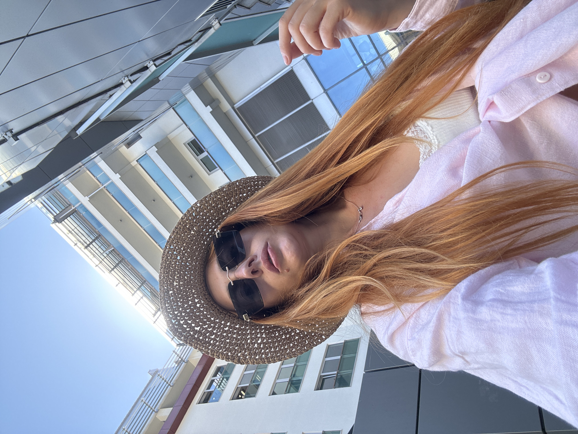 A woman with long, auburn hair is captured in a selfie-style shot, looking directly at the camera. She is wearing a wide-brimmed straw hat and dark sunglasses, obscuring her eyes. A light pink linen shirt is open, revealing a white lace-trimmed camisole underneath. She is adorned with a delicate necklace. Her right hand is visible at the edge of the frame, slightly raised.
The background features a modern building with multiple stories, characterized by a white facade, glass balconies, and silver-toned paneling. The sky above is a clear, bright blue. The overall scene suggests an outdoor setting in a city environment. The architecture and style are consistent with contemporary buildings found in Budva, Montenegro, a popular coastal city.
