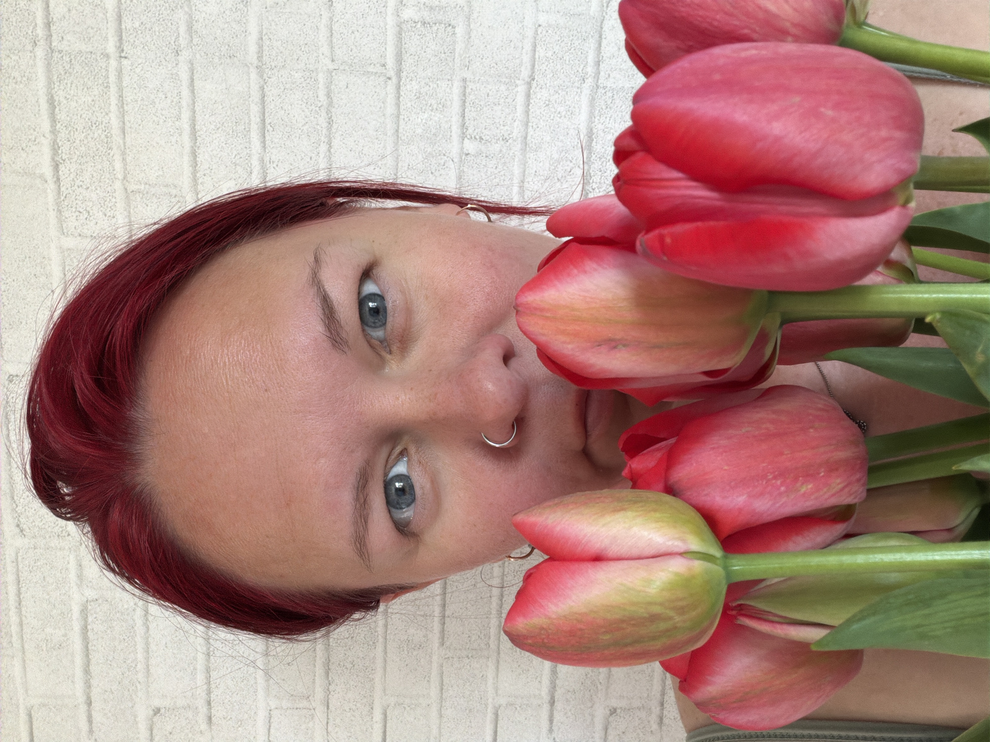 A person with red hair and blue eyes is holding a bouquet of red tulips in front of a white brick wall. The person has a nose ring. The location is unknown and there is no information on the city of Chaikovsky, Russia.
