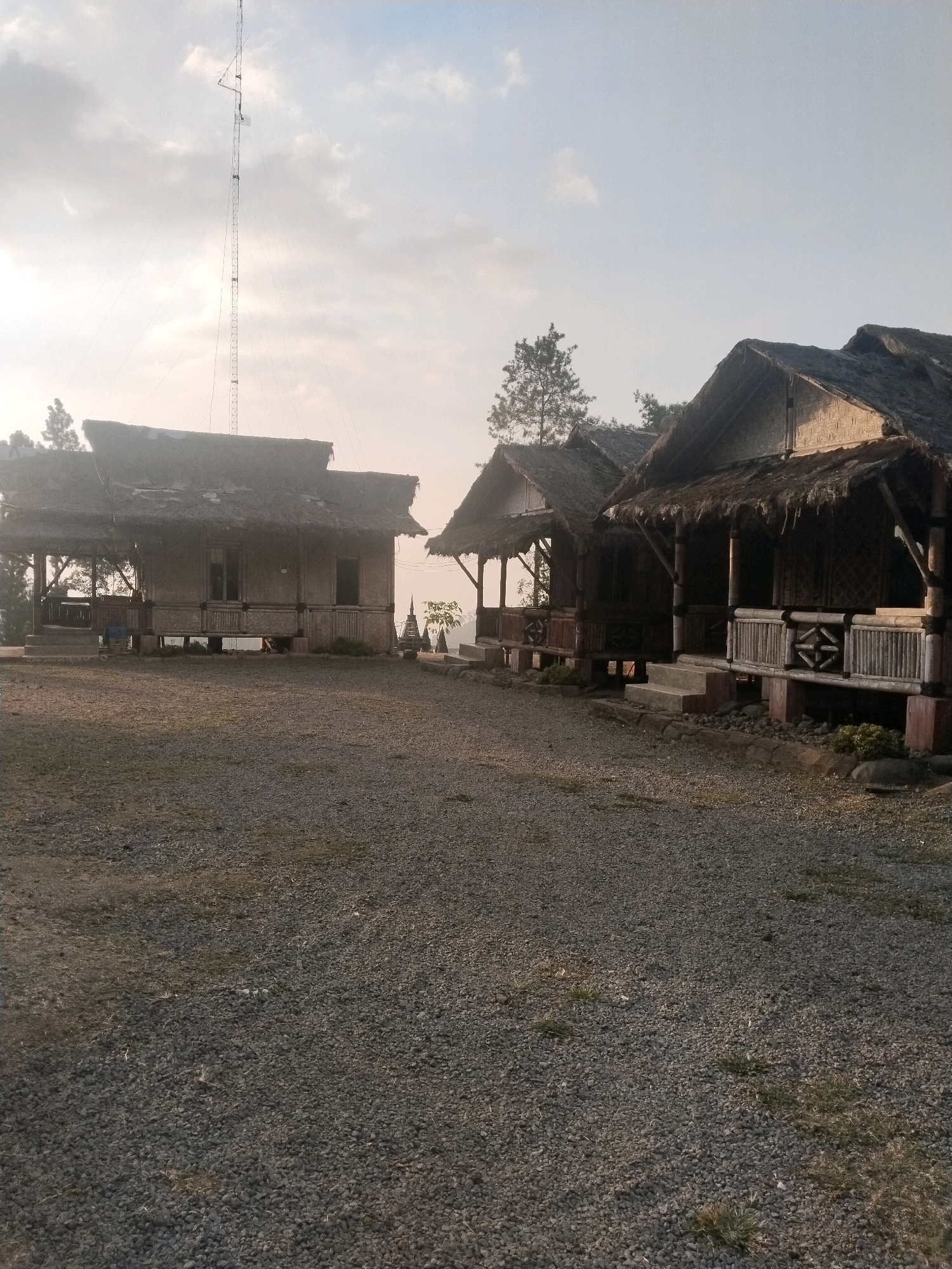 The image depicts a cluster of traditional bamboo huts with thatched roofs, likely guest accommodations, situated on a gravel-covered area. The huts are elevated on stilts, and some feature open verandas with bamboo railings. The architecture suggests a rural or resort setting, consistent with the natural beauty of Guci, Indonesia, a region known for its hot springs and scenic landscapes. In the background, a tall transmission tower stands against a hazy sky, hinting at the presence of telecommunication infrastructure within or near the vicinity. The overall impression is one of rustic simplicity and a tranquil, natural environment, possibly capturing a sunrise or sunset due to the soft, diffused light. The ground is covered with small, dark grey stones, and sparse patches of dry grass are visible. The huts themselves are constructed with woven bamboo walls and prominent, sloped roofs made of dried natural materials. Some of the huts appear to have windows and doors, but their interiors are not clearly visible. The context of Guci, Indonesia, implies this scene is likely part of a tourist destination or a community that embraces traditional building methods.