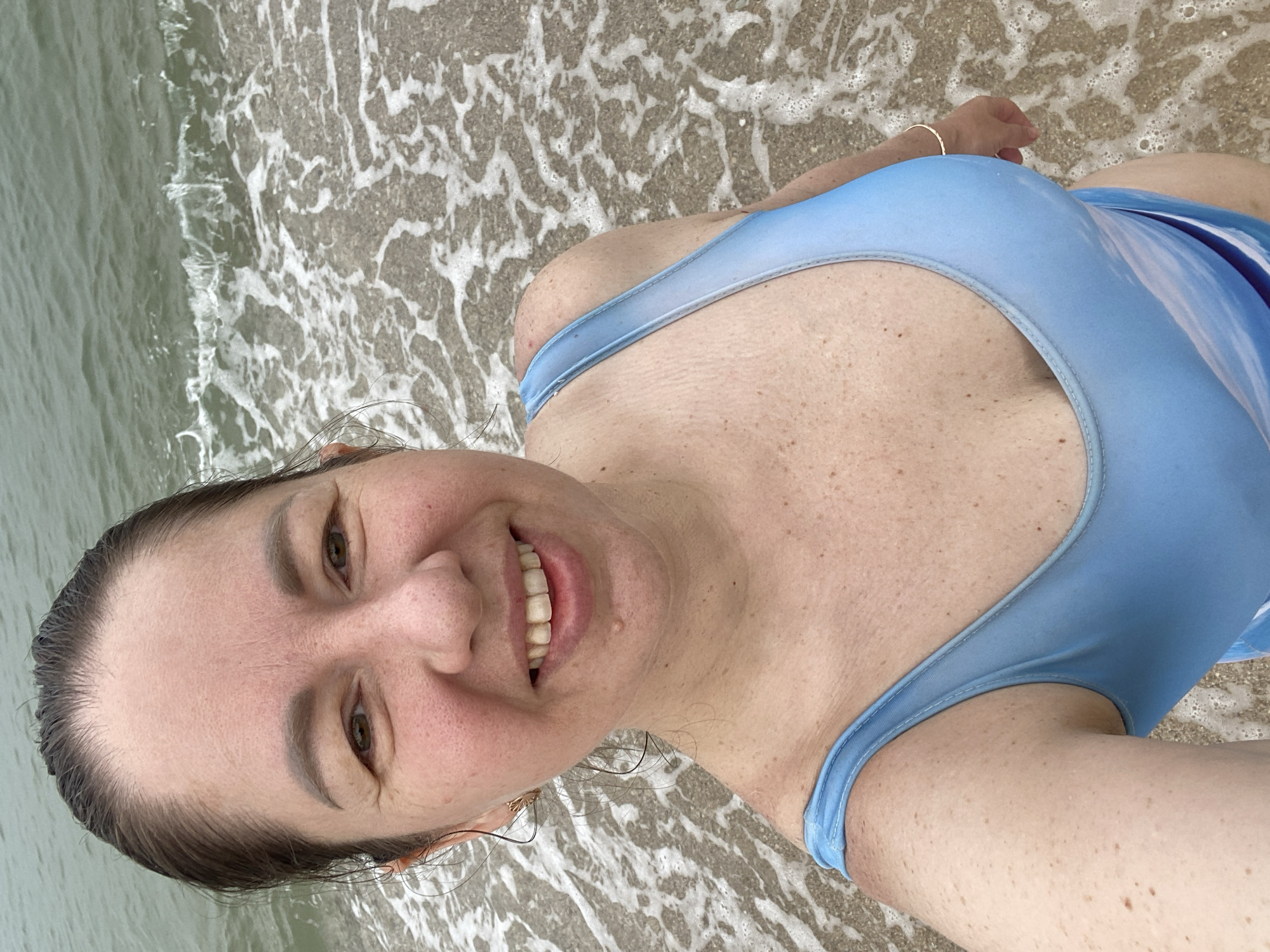 A middle-aged woman in a light blue bathing suit smiles for a selfie on a beach. Waves are lapping at the shore near her feet. The location is Phu Thuy Ward, Vietnam.