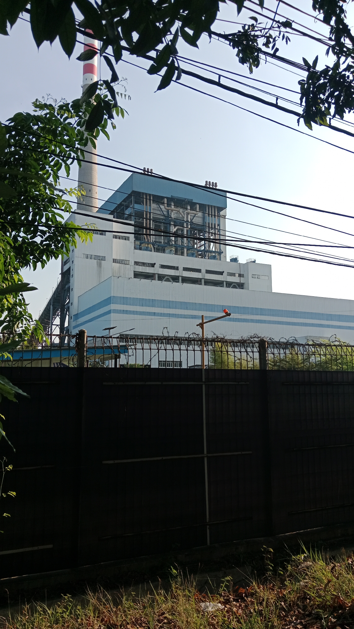A tall, white smokestack with red bands stands to the left of a large, white industrial building with a blue roof and exterior blue stripes. Power lines stretch across the sky above the building. A black fence topped with barbed wire runs in front of the building, with a red beacon on a pole to the right of the fence. Green foliage partially obscures the view of the building and smokestack. The scene is set in a dry, grassy area in front of the industrial complex.