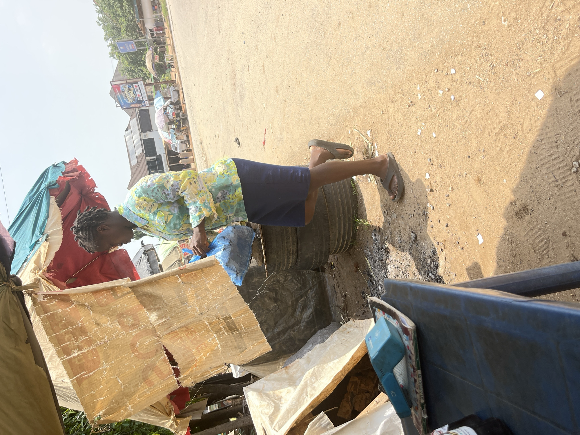 A person, likely female, is standing outdoors near a street in Abuja, Nigeria. The person is wearing a colorful patterned short-sleeved shirt and a dark blue skirt. They are also wearing dark-colored flip-flops. The person is bending over and appears to be holding a blue plastic bag. The ground is sandy and appears to be a dirt road or pathway, with some debris scattered around. In the background, there are buildings and trees. There is also a pile of tires and what appears to be makeshift shelters or stalls constructed from tarps and other materials. The lighting suggests it is daytime, likely sunny. The overall impression is of a bustling outdoor environment, possibly a market or a street with informal vendors. A blue plastic object with some text on it is visible on the right side of the image. A street sign with some text is also visible in the background.