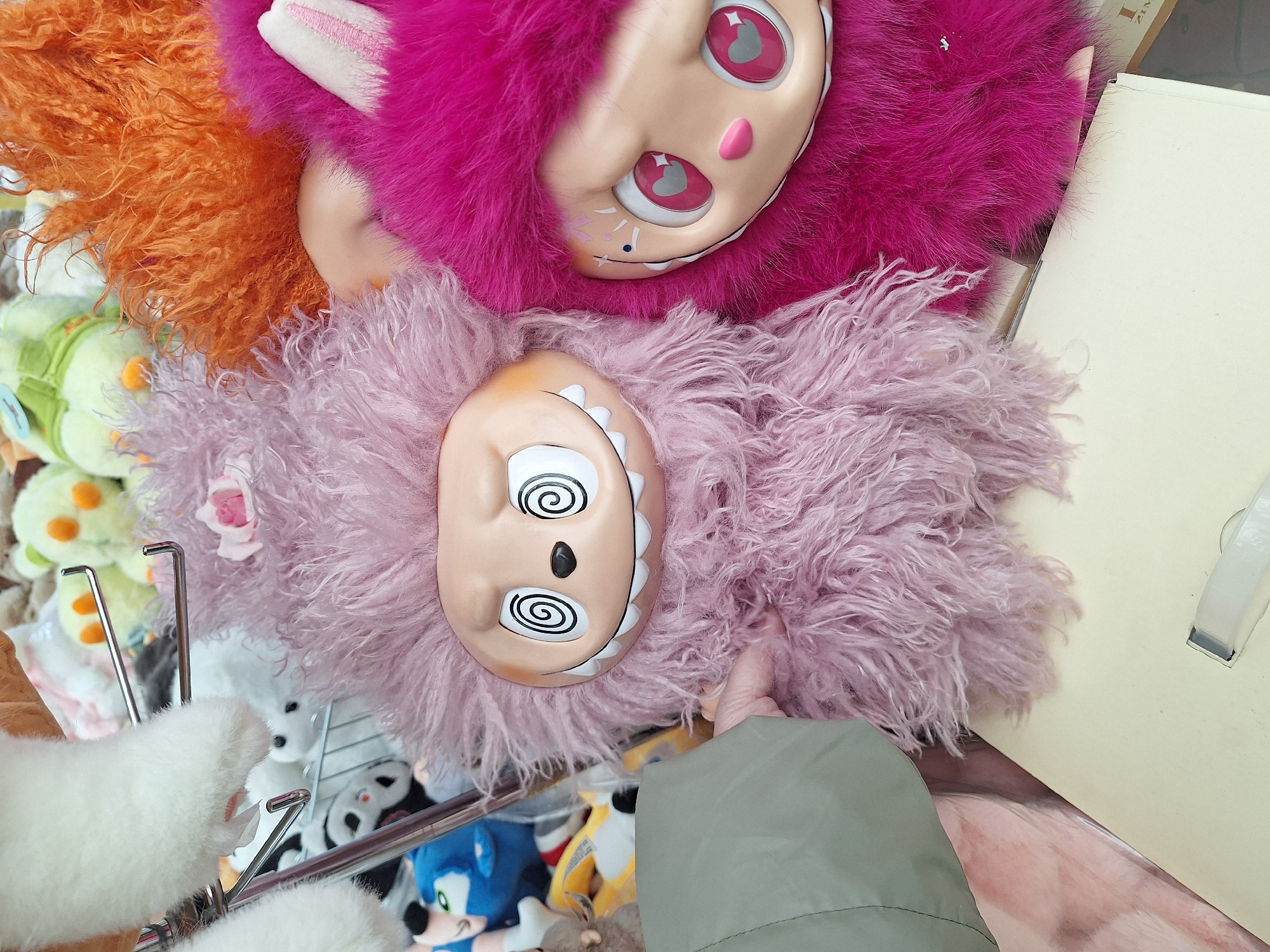 The image presents a close-up view of several plush toys. A pink plush toy with a cartoonish face and heart-shaped eyes sits atop a lilac-colored plush toy with swirled eyes. To the left of these is an orange plush toy. The scene includes other plush toys in the background, including a Sonic the Hedgehog plushie and other assorted animals, arranged on what appears to be a display rack. A sleeve of an olive green garment is visible in the foreground.