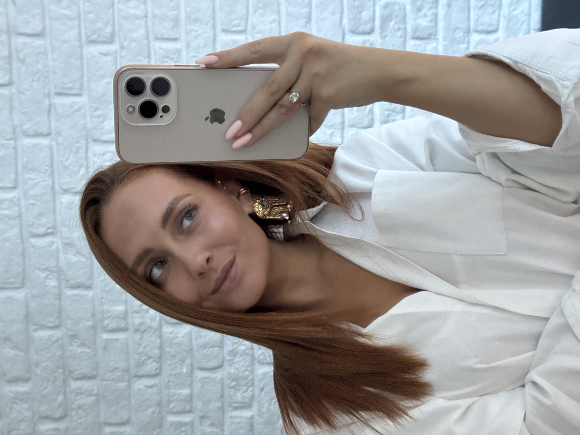 A young woman with long auburn hair takes a selfie in front of a white brick wall. She is wearing a white collared shirt, a pair of gold earrings, and a ring on her left hand. Her nails are painted a light pink. She is holding a smartphone in her right hand, which is angled to capture her reflection. The phone is in a rose gold case, and the Apple logo is visible on the back. The woman is looking to her left, with a slight smile on her face.