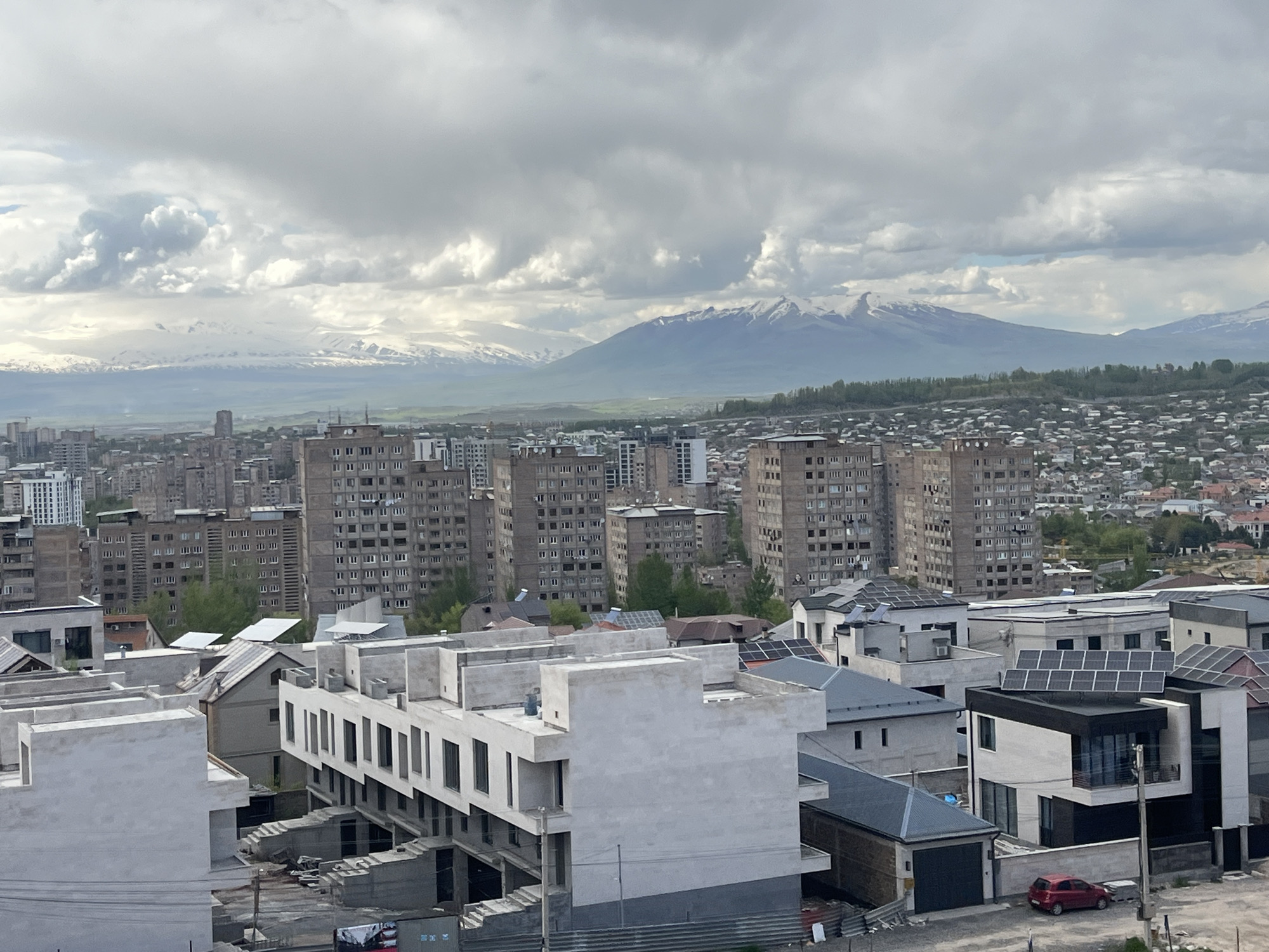 The image captures a cloudy day in Arinj, Armenia, featuring a mix of old Soviet-era apartment blocks and new residential construction. In the foreground are newly built homes, some still under construction, with modern architectural styles and solar panels on some roofs. A red car is parked near one of these new houses. In the mid-ground, older, taller apartment buildings dominate the cityscape. Snow-capped mountains are visible in the distance, partially obscured by clouds. The overall environment conveys a blend of urban development and natural landscape under an overcast sky.