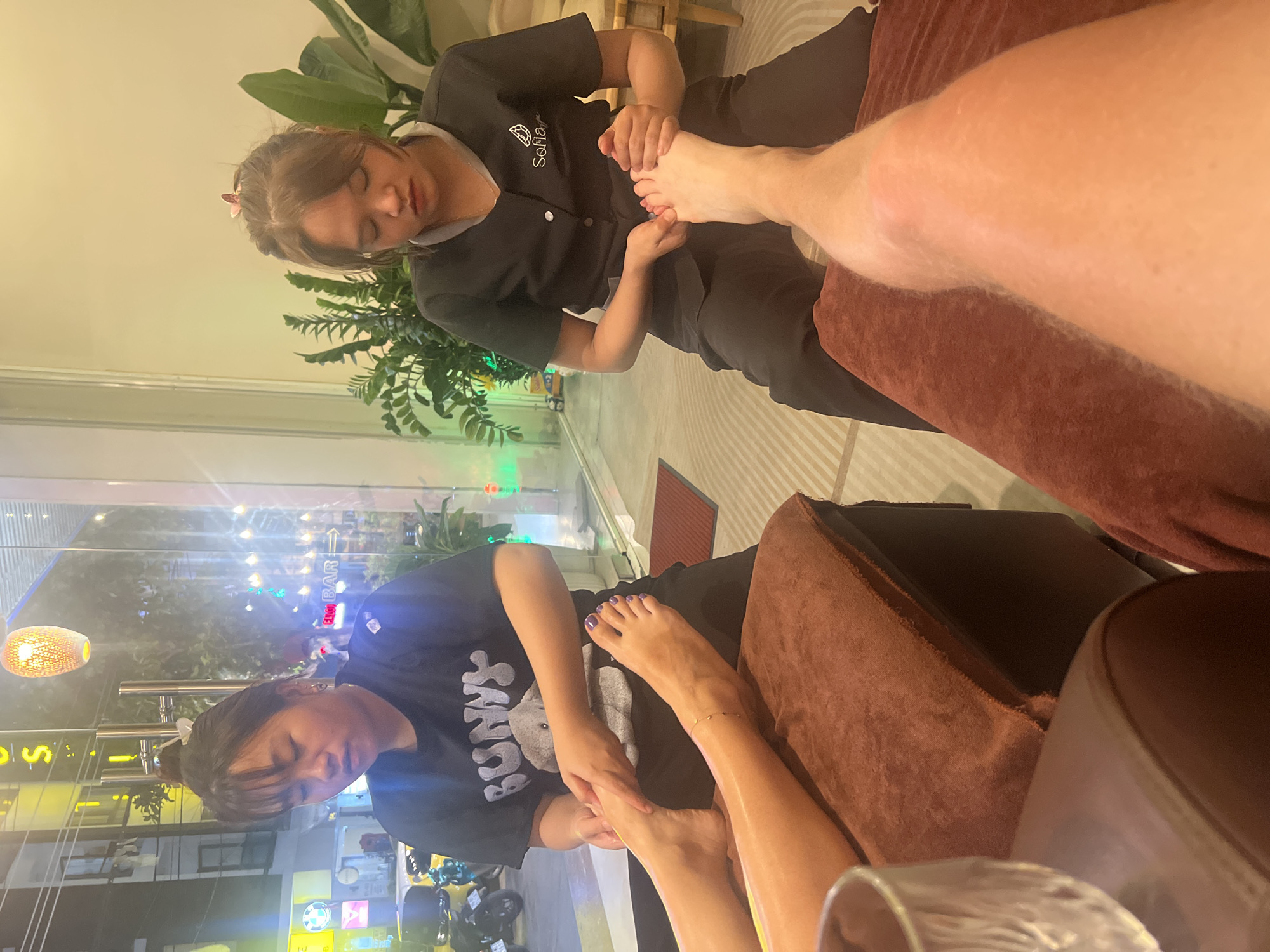 The image shows two people receiving foot massages indoors in an establishment located in an unidentified city. In the upper half of the image, a woman wearing a black uniform with a logo on the upper right side is giving a foot massage to a person with fair skin. A plant is visible in the background. In the lower half of the image, another woman in a black t-shirt is giving a foot massage to a person with purple painted toenails. The view through the window shows a street scene with motorbikes and storefronts, suggesting a commercial area. The lighting is a mix of natural light and artificial light, creating a warm ambiance. The focus is on the foot massages, with the feet and the hands of the masseuses being the prominent elements in the composition.
