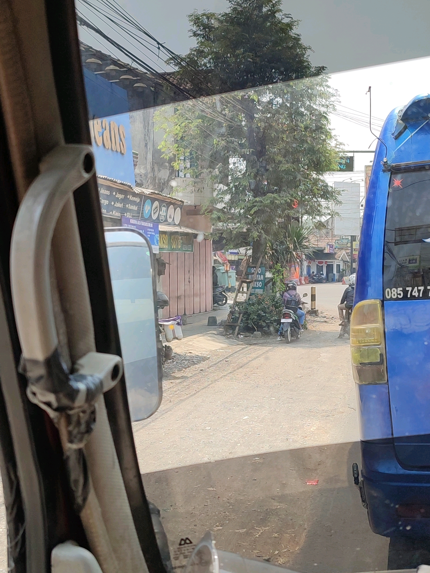 The image shows a street scene in Secang, Indonesia, viewed from inside a vehicle. In the foreground, a part of the vehicle's interior is visible, including a grey grab handle secured with black tape. To the right, the rear quarter panel of a blue bus is prominent, with a visible license plate reading "085 747 7". The bus is parked or moving slowly on a dusty road.
Across the street, several shops are visible. One shop has a blue facade with large yellow lettering that appears to spell "JEANS". Below this, a sign lists various clothing items like "Jeans", "Joger", "Kemeja", "Cargo", "Jaket", and "Sandal". Further down the street, a tree provides shade, and several motorcycles are parked or in motion. Two people are riding motorcycles on the street. One person on a scooter is wearing a helmet and a purple jacket, while another person on a motorcycle is wearing a helmet and a dark jacket. The buildings in the background suggest a commercial area. The environment appears to be sunny with some shadows cast by the trees and buildings. Electrical wires are strung overhead.
