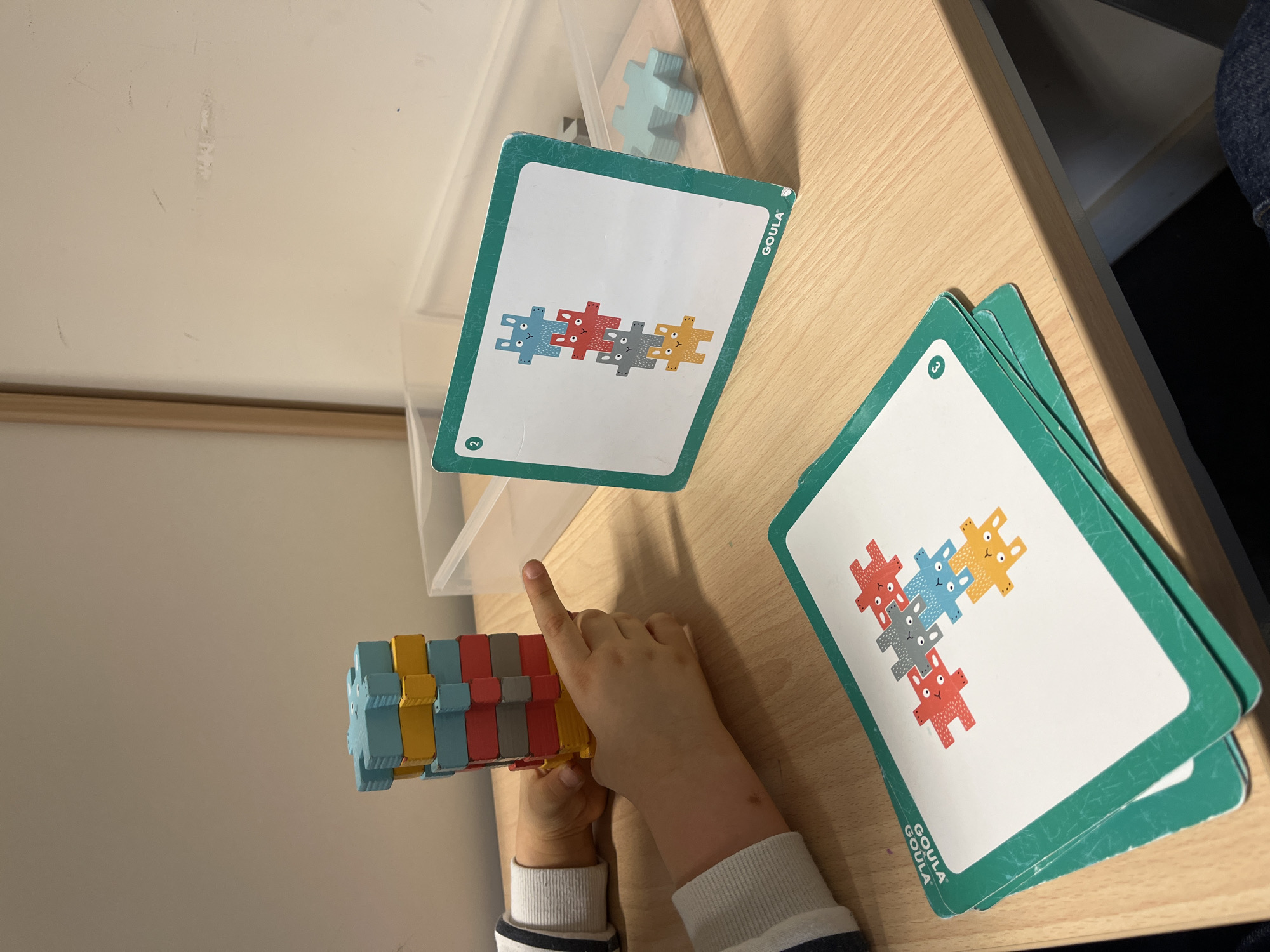 A child is playing with a Goula wooden puzzle on a light wood-grain table. The child is holding a partially constructed puzzle of interlocked colored pieces (light blue, yellow, red, gray) in their hands, seemingly matching a pattern on a Goula card in front of them. There are several Goula cards stacked nearby, with the top card displaying a pattern of red, gray, light blue, yellow, and red pieces. The puzzle pieces are being held in a container. The location is possibly La Fouillouse, France, suggested by "Goula" being a Spanish toy brand, but the environment depicted does not provide definitive proof of this location. The child's hands and forearms are visible, clad in a long-sleeved garment with a striped cuff. The background includes a light-colored wall.