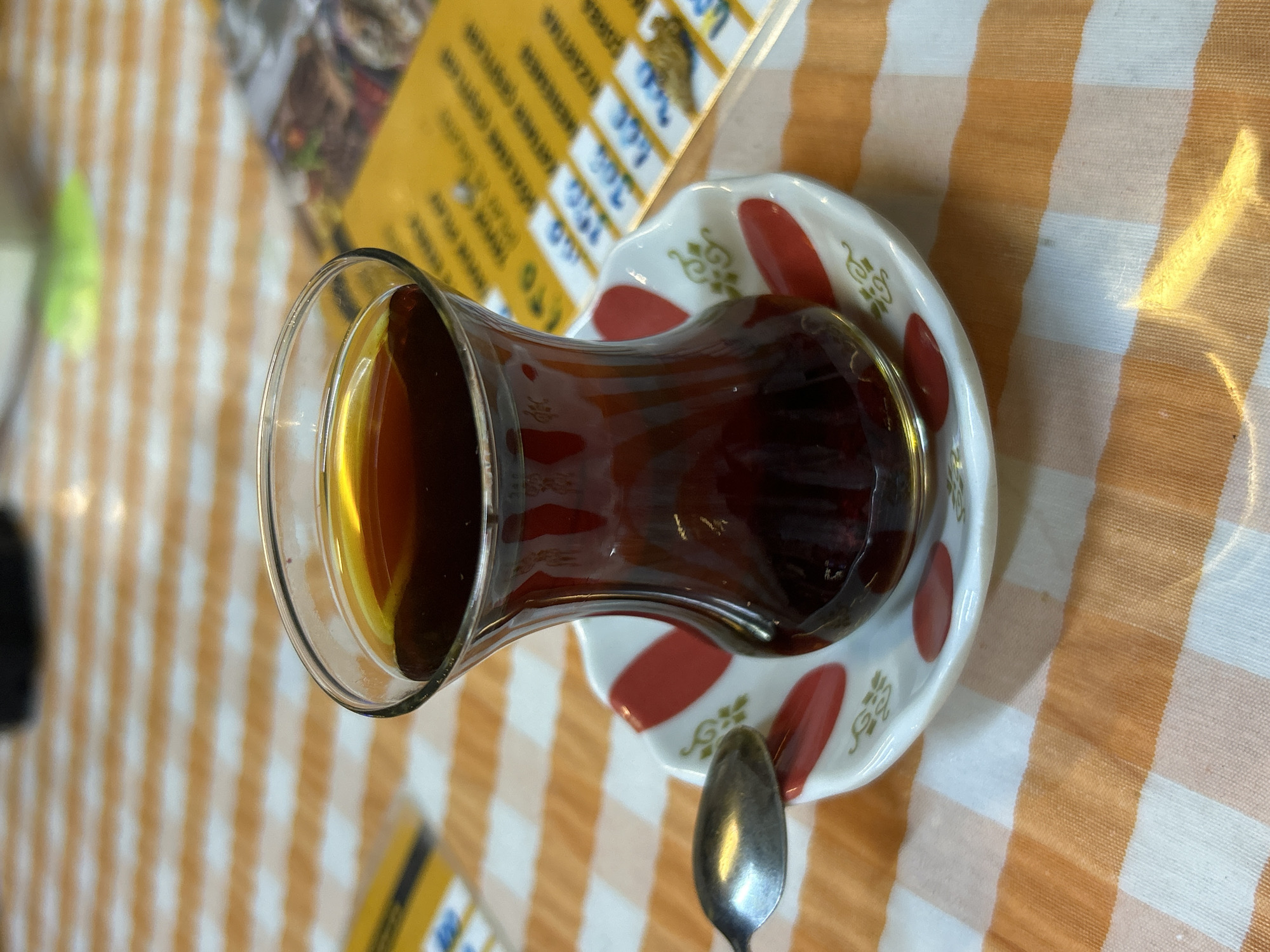 A close-up, slightly elevated shot shows a glass of dark amber tea resting on a saucer on a checkered tablecloth. The glass is tulip-shaped and filled to about three-quarters capacity. The saucer is white with red oval accents and gold scrollwork. A silver spoon rests on the saucer to the left of the glass. In the background, out of focus, is a yellow menu with Turkish text and some food images. The scene is set in a cafe or restaurant in Ortaca, Turkey, suggested by the presence of a menu with Turkish text. The lighting is warm and seems to come from above, creating highlights on the glass and spoon.
