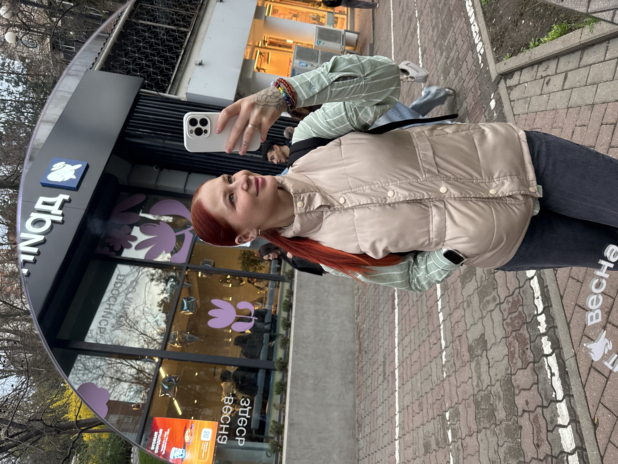 A woman is taking a selfie outside a building in Ovoshchnoy, Russia. The building has a sign in Russian with an image of a rabbit. The window displays purple flower decorations, and signage indicates "Здесь весна" (Here is Spring). The woman wears a light tan vest over a striped shirt, jeans, and bracelets. She is holding a white smartphone. The surrounding environment shows a sidewalk with parking stripes and additional text in Russian.