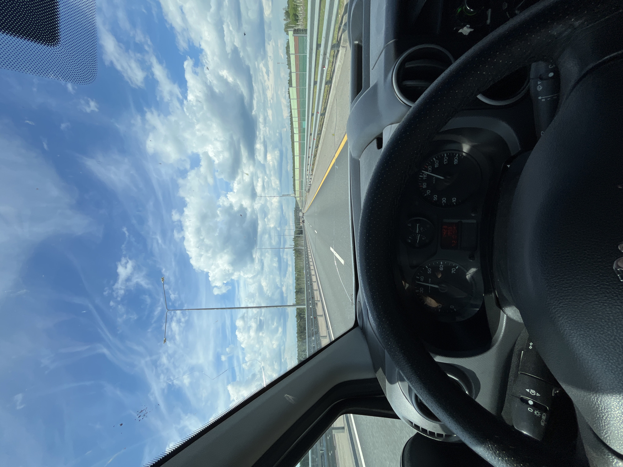 The image presents a driver's perspective from inside a vehicle traveling on a highway, likely near Вельмогово, Russia, during daytime. The prominent subjects include the car's interior on the right, and the open road stretching ahead under a vast, bright blue sky filled with fluffy white cumulus clouds and wispy cirrus clouds.

The scene depicts driving on a multi-lane asphalt highway. To the right, a long sound barrier or guard rail, featuring alternating green and orange vertical panels, runs alongside the road. Beyond this barrier, a dense line of trees indicates a forested area. Several street lights on tall poles are visible along the left side of the road, extending into the distance. The windshield shows minor specks and dust.

Inside the vehicle, a black steering wheel with a textured grip dominates the right side of the frame. Part of the dashboard, including an air vent, is visible. The speedometer dial is clear, indicating speeds in kilometers per hour with markings at 0, 10, 20, 30, 40, 50, 60, 70, 90, 110, 130, 150, 170, and 190. A digital display below the speedometer shows "00" and other unreadable symbols. A control stalk, likely for wipers and indicators, is visible to the right of the steering wheel, showing light and wiper symbols. The sunny weather and clear sky suggest pleasant driving conditions.