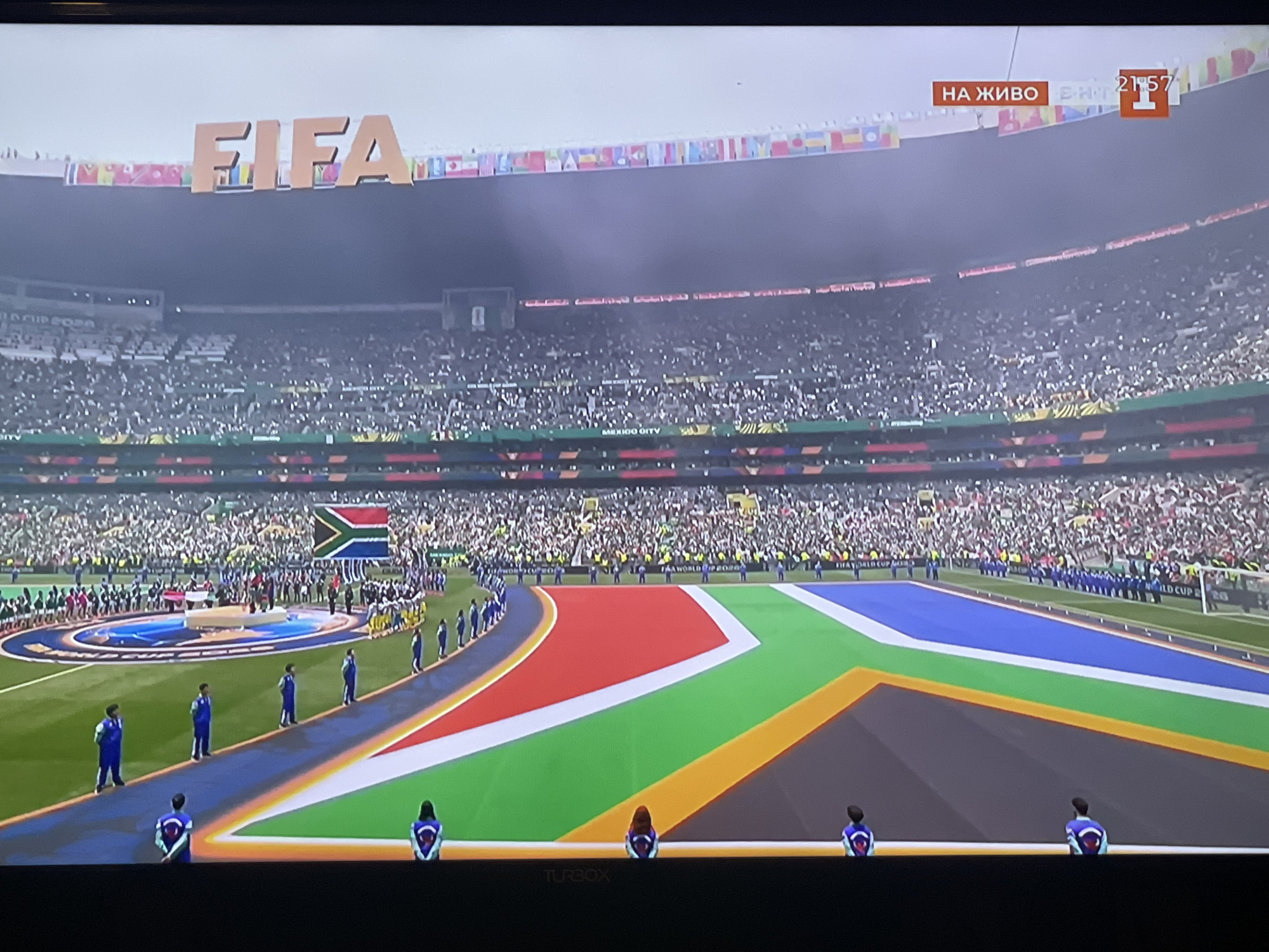 The image captures a wide view of a football stadium during what appears to be a pre-match ceremony. In the background, the stands are filled with a large crowd of spectators. Above the stadium, large, illuminated letters spell out "FIFA". A banner displaying flags of various nations stretches across the top of the stadium structure. A scoreboard in the upper right corner shows "НА ЖИВО" (meaning "LIVE" in Bulgarian) and a time of "21:57" with a FIFA logo.
In the foreground, the field is marked with vibrant colors forming a large pattern, reminiscent of a country's flag, with prominent sections of red, green, blue, and white. The South African flag is displayed prominently in the middle ground. A line of individuals, some wearing blue tracksuits, are positioned along a path leading onto the field. Several individuals, viewed from behind, are also standing on the field, wearing jackets with the South African flag motif on the back. The overall atmosphere suggests a significant sporting event. There is no visual information in the image that directly links this scene to the city of Berkovitsa, Bulgaria.