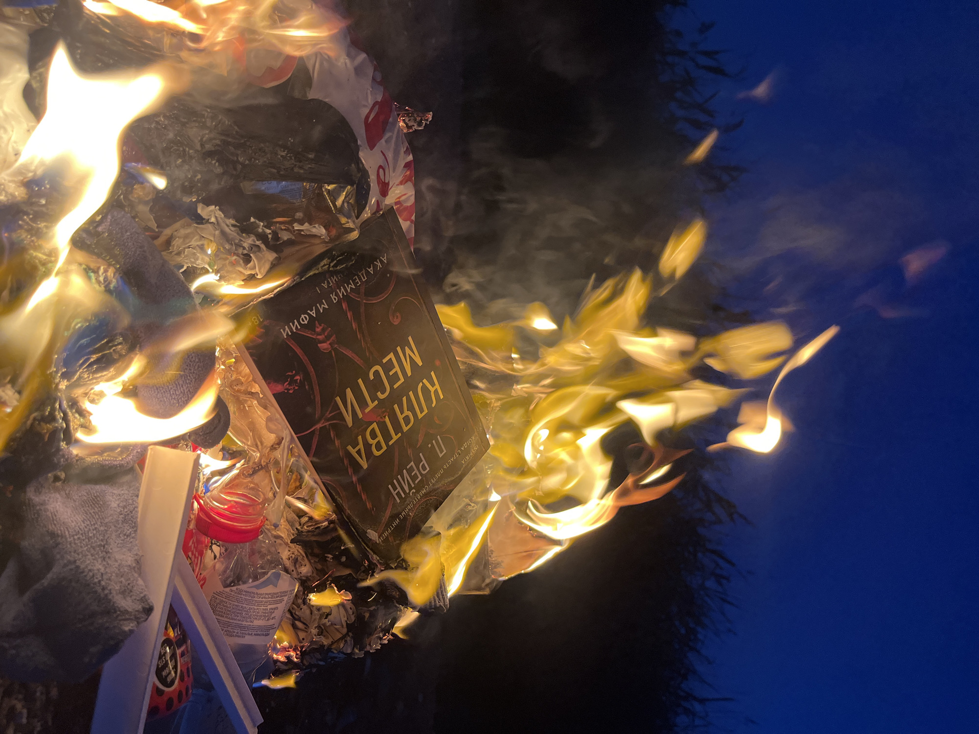 The image shows a pile of trash being burned, with a paperback book titled "Клятва Мести" (Oath of Revenge) by P. Рейн (P. Reyn), part of the "Академия Мафии" (Mafia Academy) series, also being consumed by the flames. The book is a Russian language edition. Among the trash are various items including a plastic bottle with a red cap and a grey cloth-like object. The fire is emitting smoke and flames, casting light onto a dark background, which appears to be night sky, due to its blue color. The image does not contain details about Kaspiysk, Russia. There are no people depicted in the image.