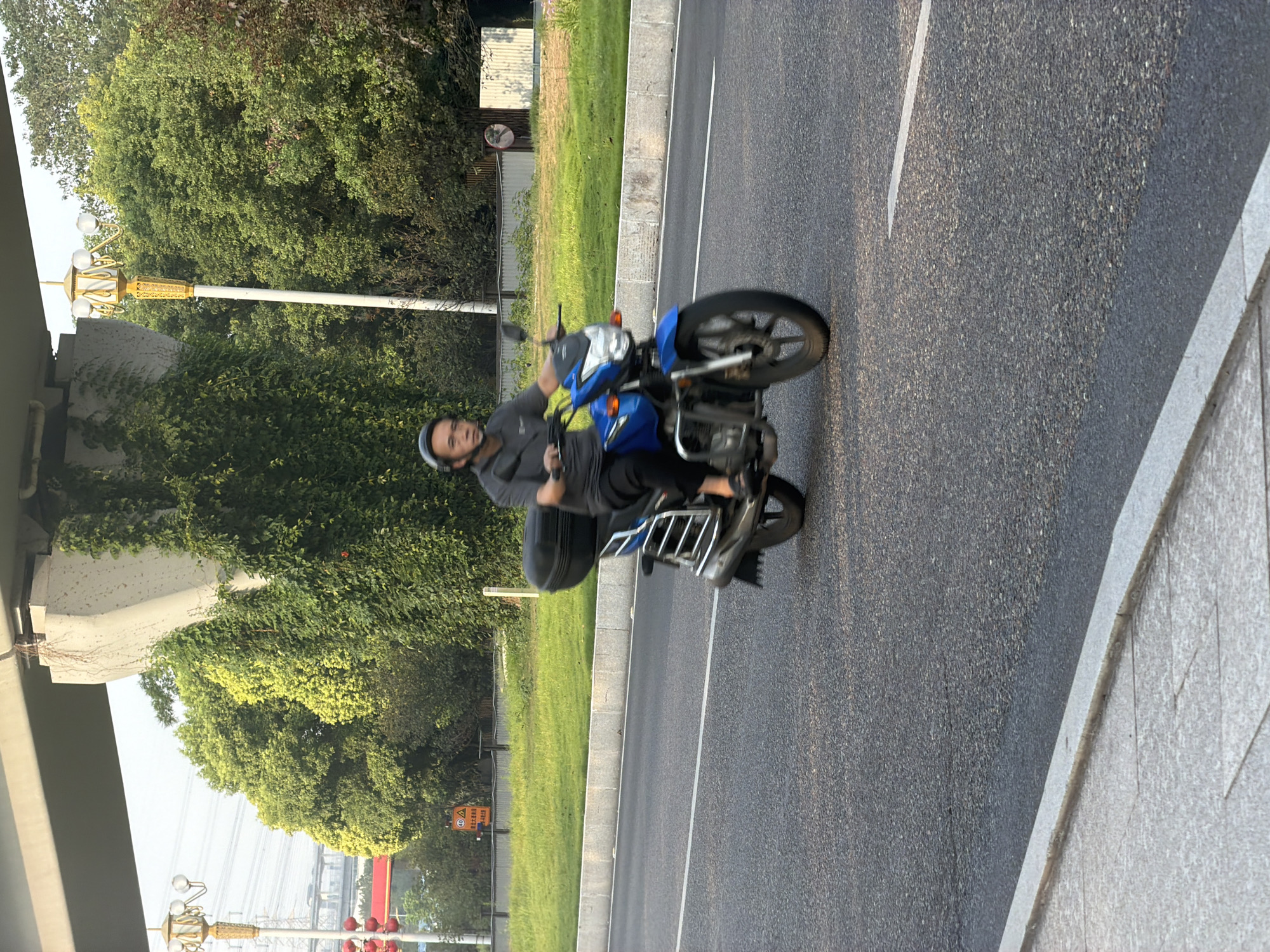 A man wearing a helmet rides a blue motorcycle with a black storage box on the back on a road in Jiangjin District, Chongqing, China. The man is wearing a grey shirt and dark pants. The motorcycle has a chrome luggage rack. The road is asphalt with a white lane marking. In the background, there is a grassy area, trees, a concrete overpass support, and a lamppost. Power lines and traffic lights are visible in the distance. The sky is overcast.
