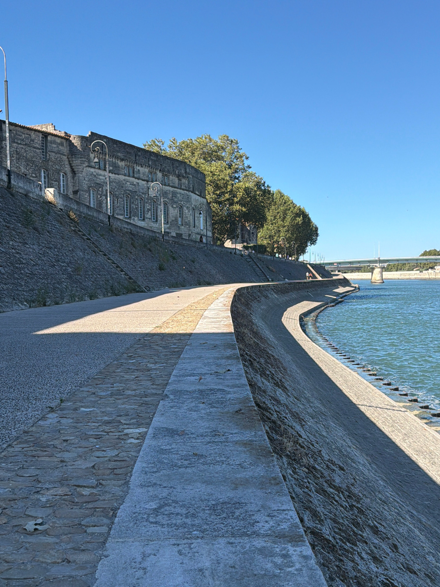 The image depicts a view along a curved riverside promenade in Arles, France, likely the banks of the Rhône River. A stone embankment lines the water, featuring steps leading down to the river in places. A paved pathway runs parallel to the embankment, with a section of cobblestones visible. Sunlight casts a strong shadow across the pathway. To the left, a steep, stone-covered slope rises, topped by a historic stone building with numerous windows. Trees line the embankment behind the building. In the distance, a bridge crosses the river. Two people are visible walking on the promenade near the bridge. The sky is clear and blue, indicating a sunny day.